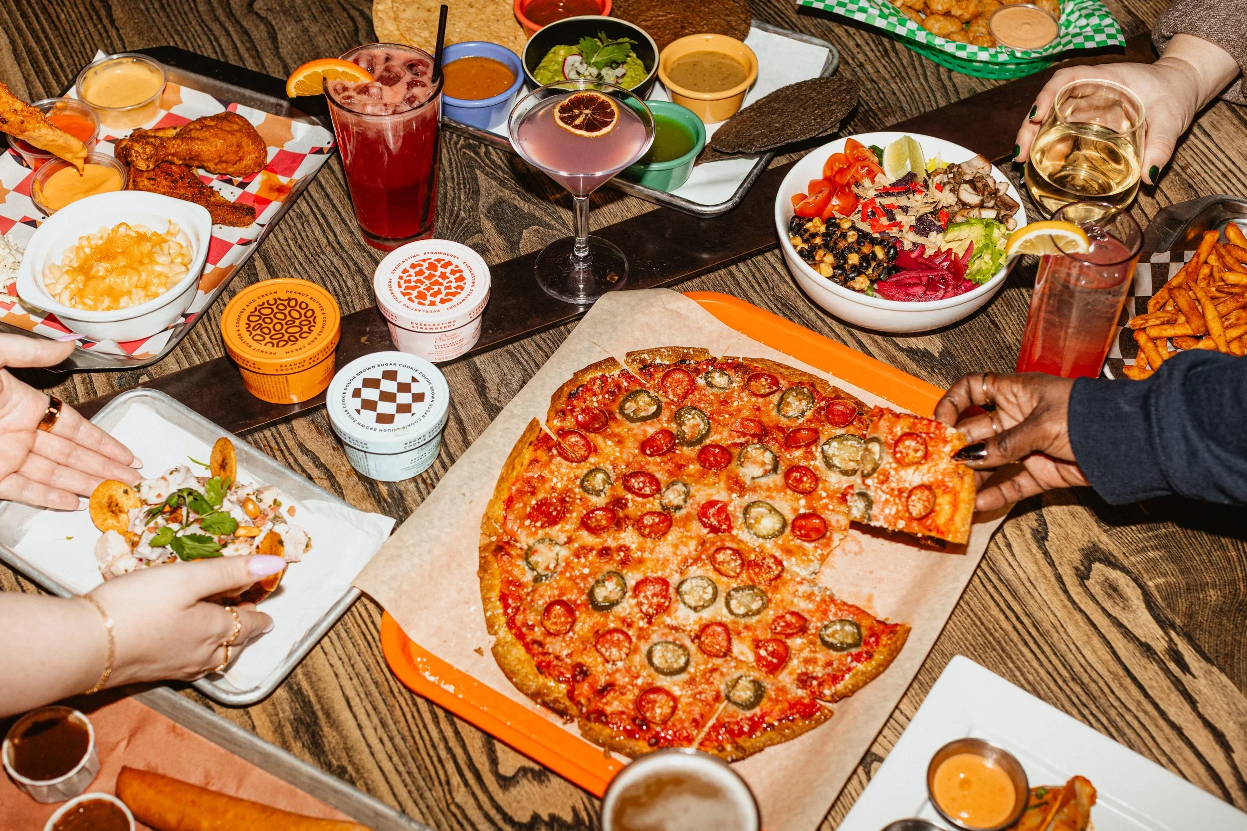 A table filled with various dishes including a pepperoni and jalapeño pizza, fried chicken wings, salads, fries, and assorted dips and drinks at a casual dining setting.