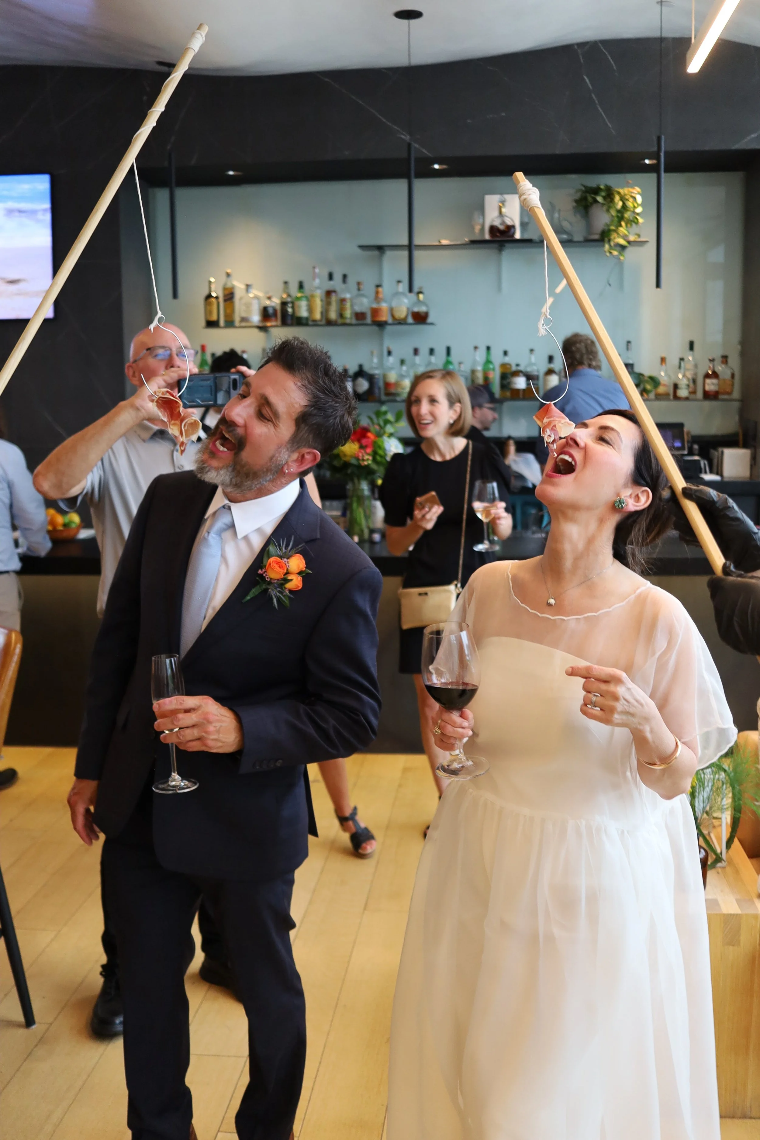 A wedding reception with a bride and groom playing a humorous game where they are biting slices of prosciutto hanging from sticks, while guests in the background smile and enjoy drinks.