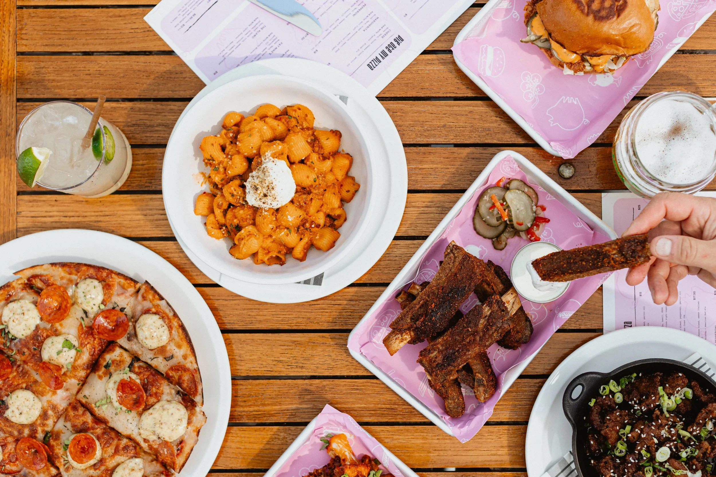 A variety of food on a wooden table includes a pizza, pasta, an assortment of ribs, pickles, a sandwich, and drinks.