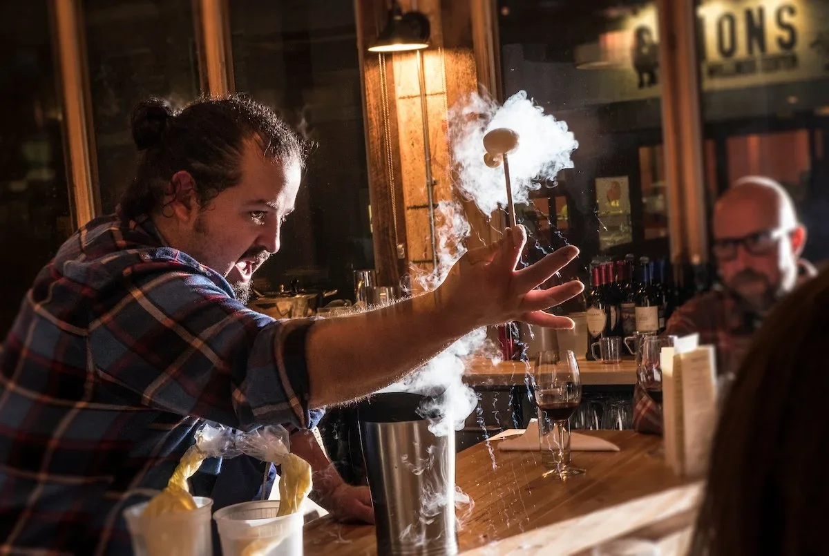 Bartender in a plaid shirt performing a smoke trick with a skewer in a bar or restaurant setting, with other patrons watching.