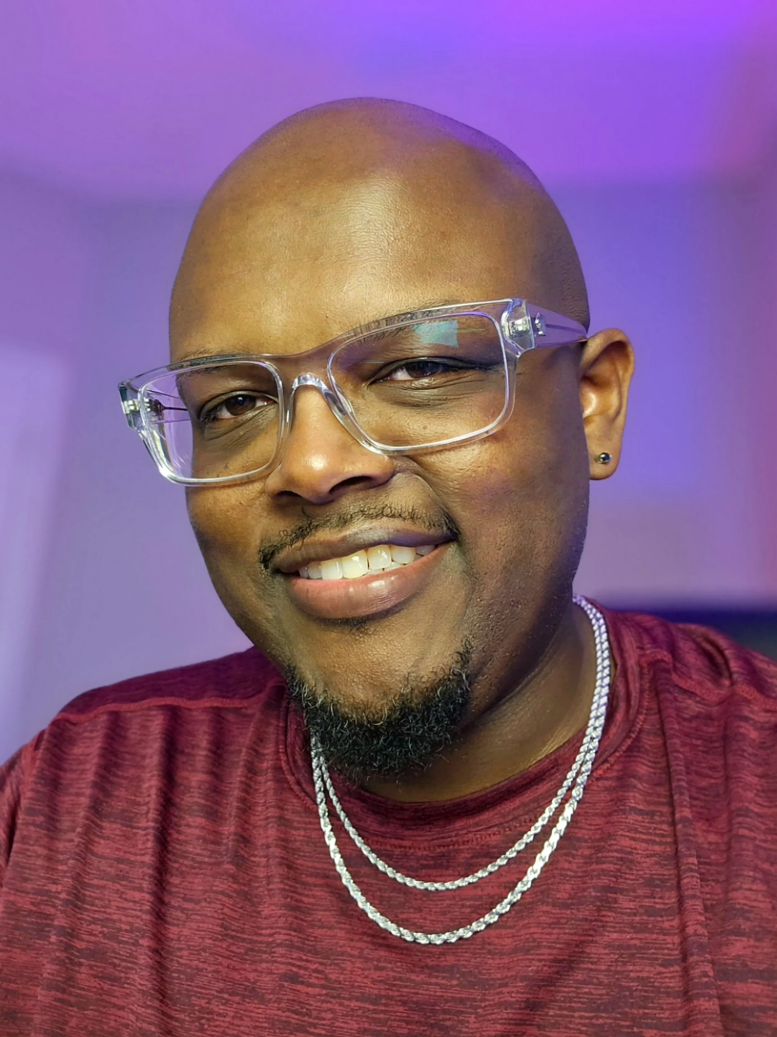 A smiling man with glasses, wearing a red shirt and layered silver necklaces, with a purple background.