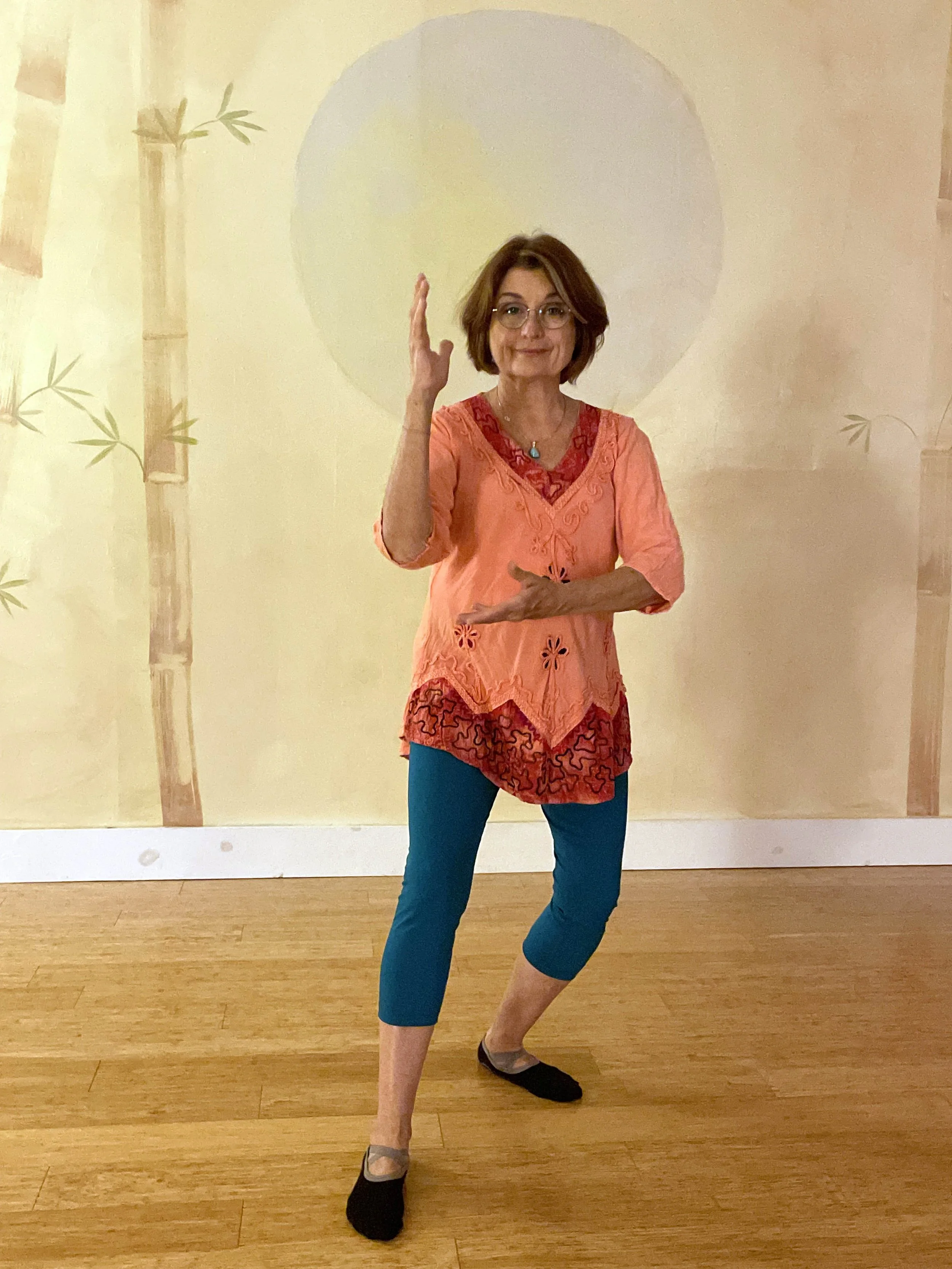 Tai Chi instructor, LinDel Sandlin mid tai chi sequence with one arm raised and one bent at her waist wearing blue leggins and a peach embroidered top