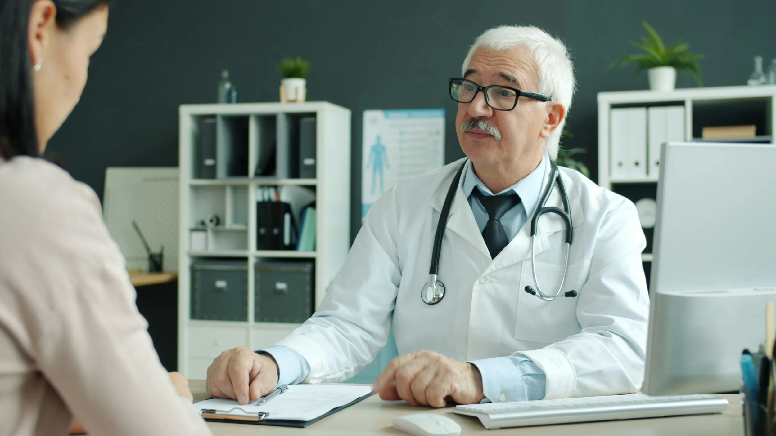 A doctor with white hair, glasses, and a stethoscope talking to a patient in an office.
