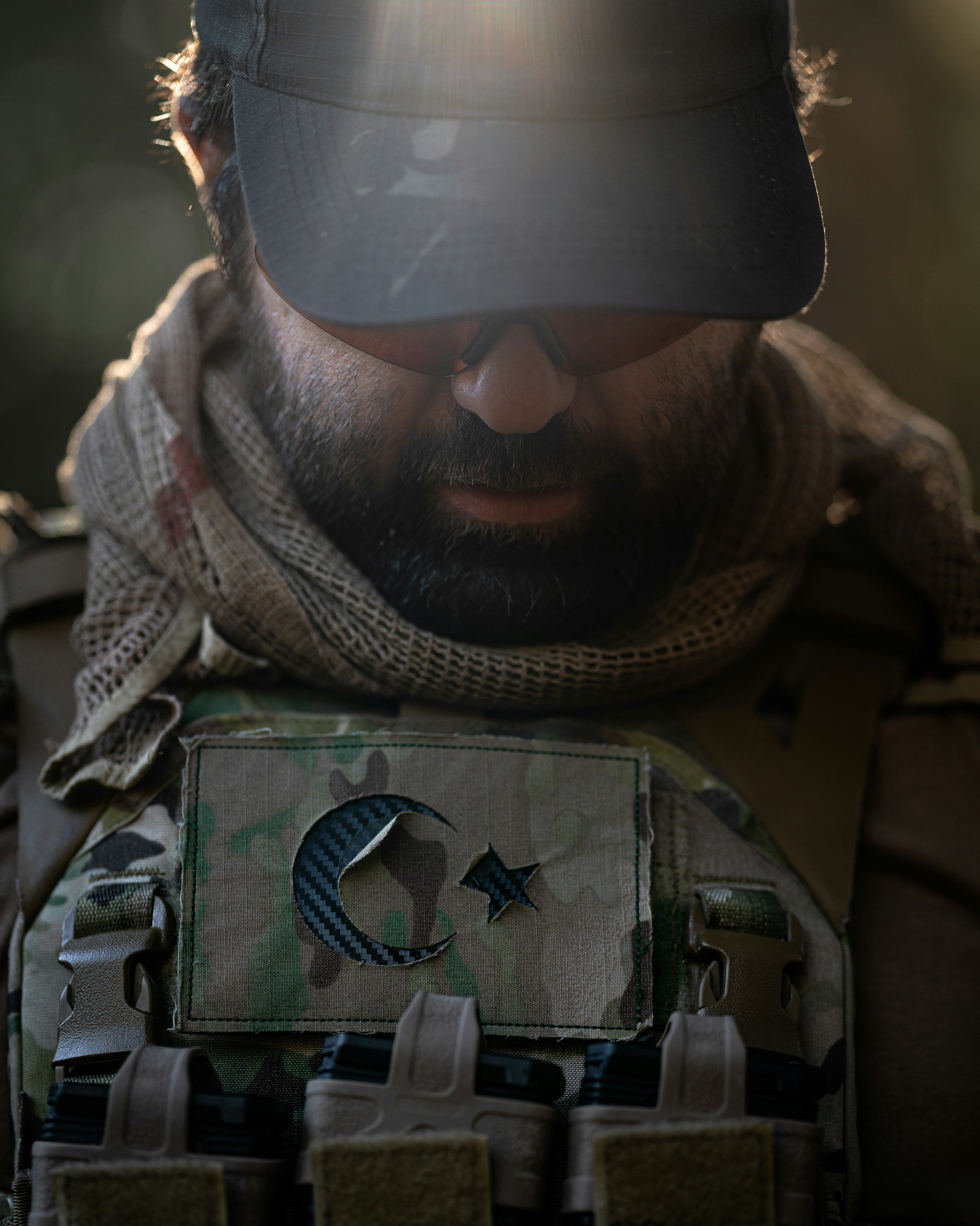 Close-up of a man wearing a black cap, sunglasses, camouflage gear, and a tan scarf with a patch featuring a moon and star, suggesting a military or tactical setting.