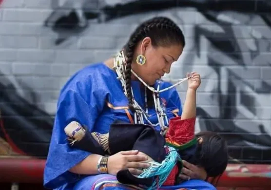 A woman dressed in blue traditional clothing with jewelry, sitting against a graffiti-covered brick wall, holding a child in her lap, and the child is playing with a necklace.