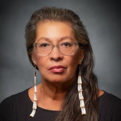 A woman with long dark hair smiling, standing against a wooden wall indoors.