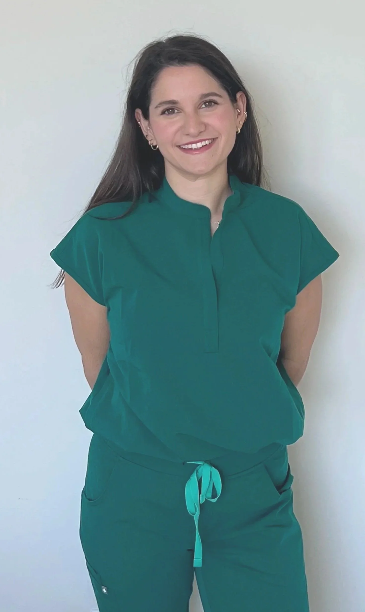 A woman with dark hair wearing a green medical scrub top and matching drawstring pants, standing against a plain white wall, smiling at the camera.
