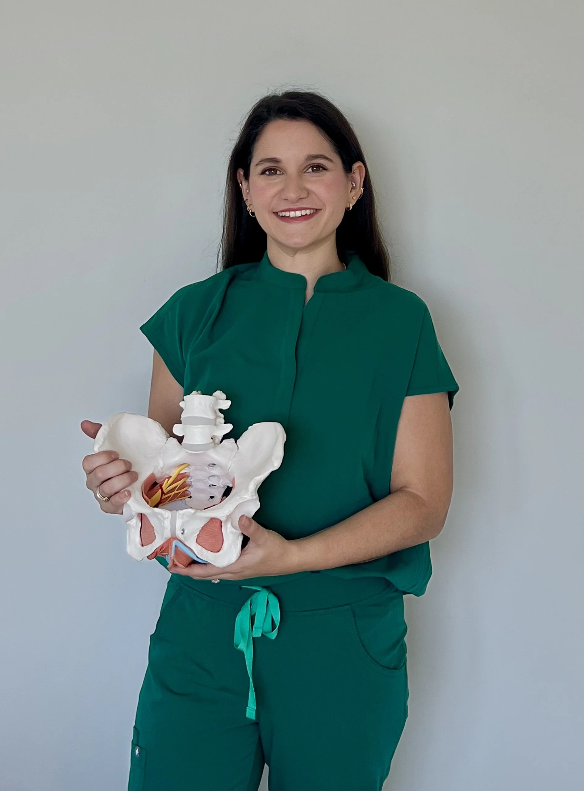 A woman dressed in green scrubs holding a model of a human pelvis and lower spine.