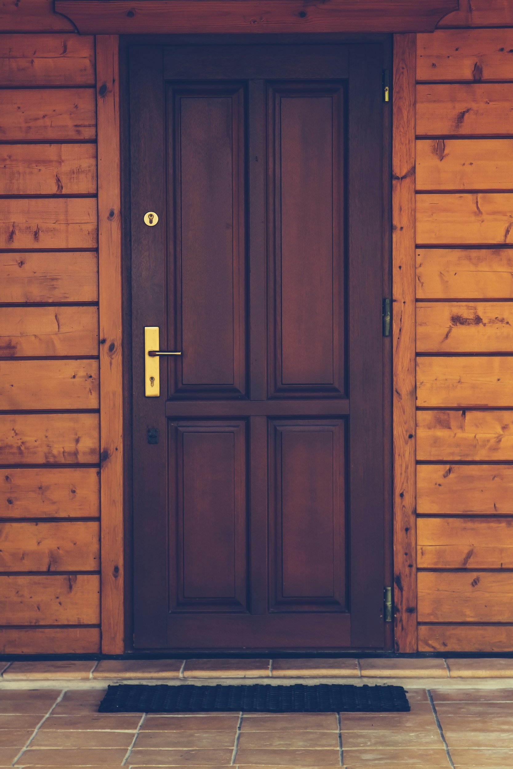 A wooden front door with a brass handle and lock, set within a wooden wall with horizontal planks, and a black doormat on a tiled porch.