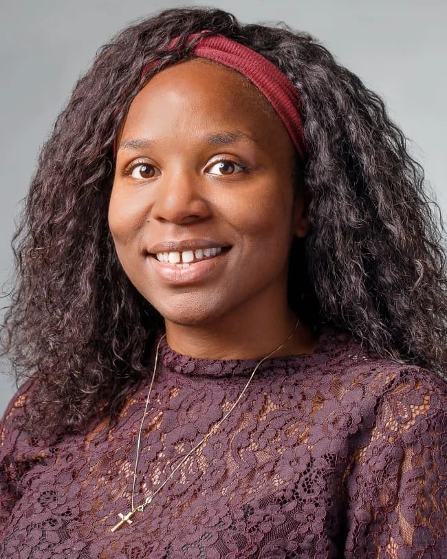 A woman with curly hair and a red headband wearing a purple lace top smiling at the camera.