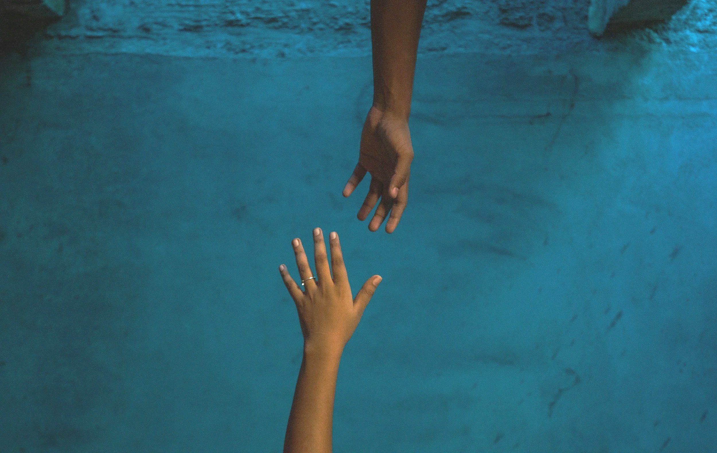 Two hands reaching towards each other, with one hand above the water and the other hand beneath the water, in a pool or body of water.