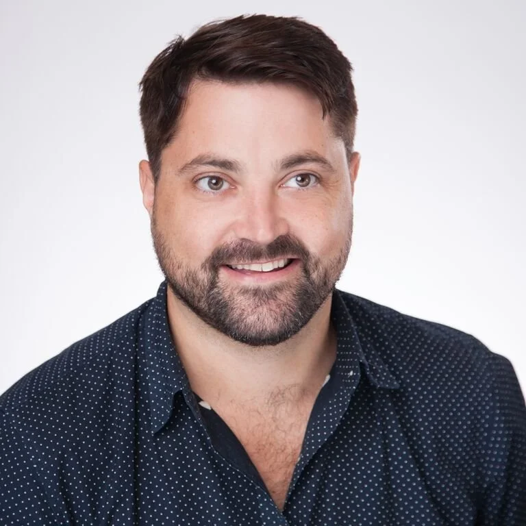 Headshot of a smiling man with short dark hair and a beard, wearing a dark blue polka dot shirt against a plain white background.
