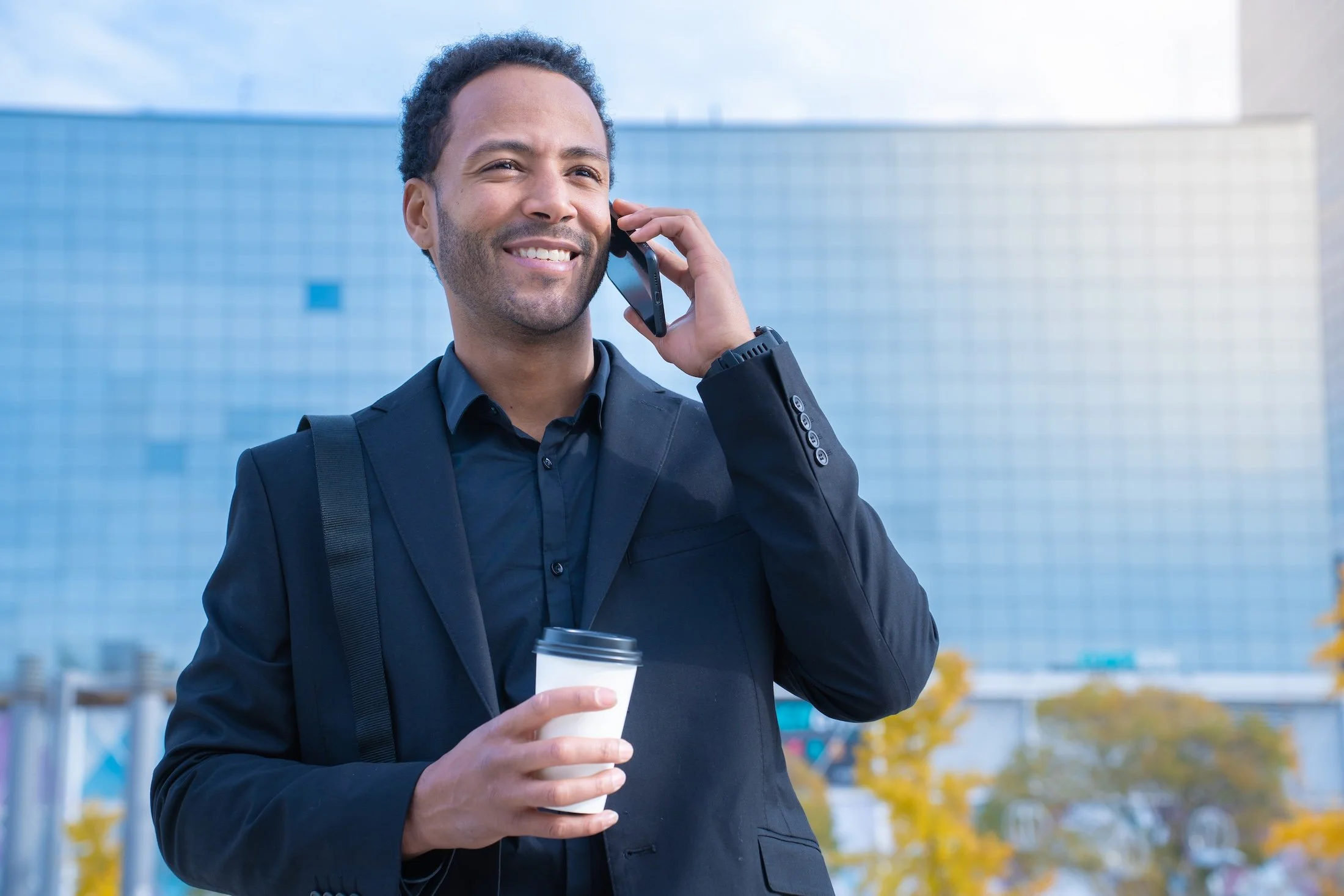 A man in a black suit holding a coffee cup and talking on his cell phone outside, with a modern glass building and autumn trees in the background.