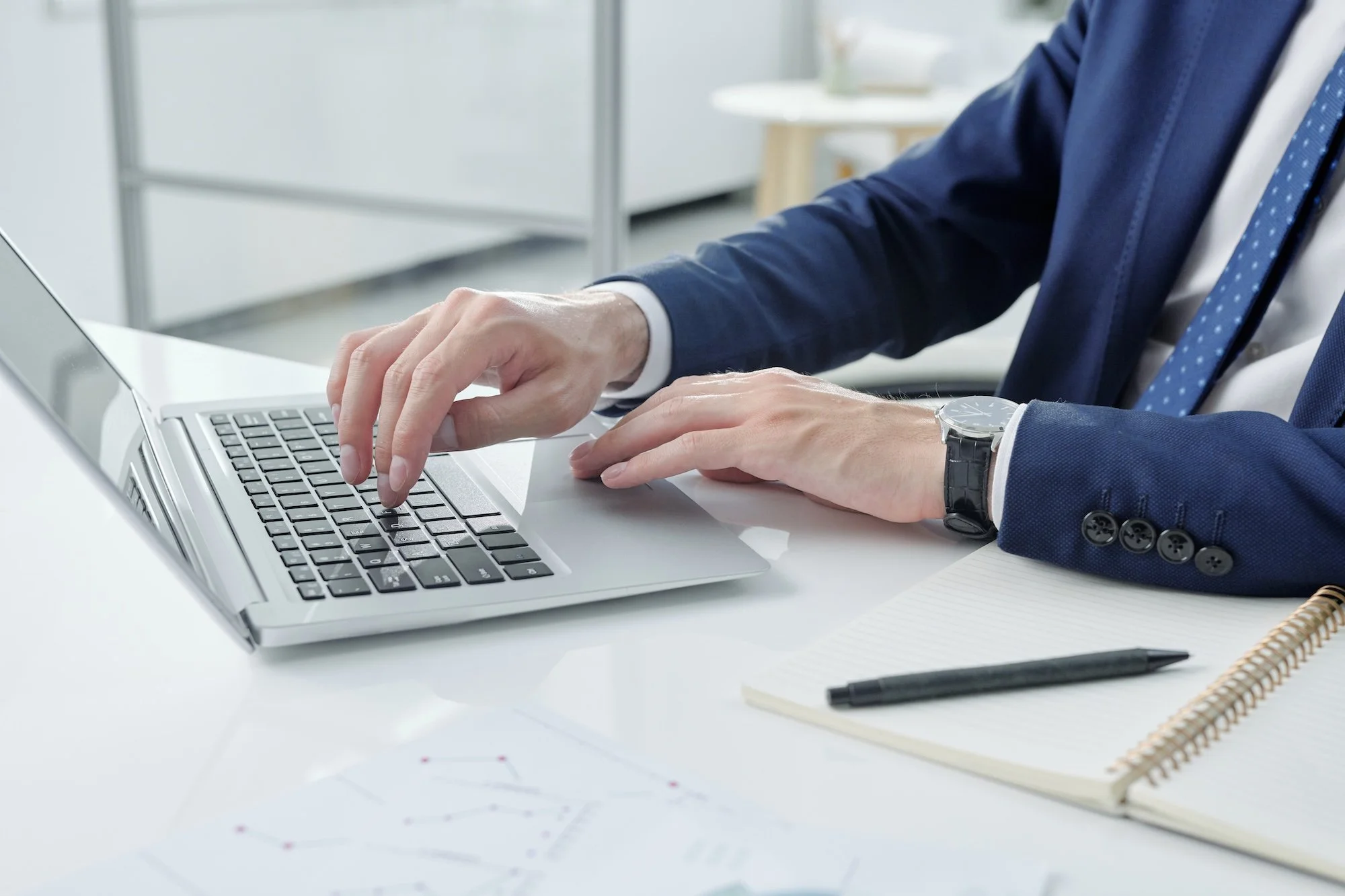 A person in business attire typing on a laptop at a desk with a notebook and pen.