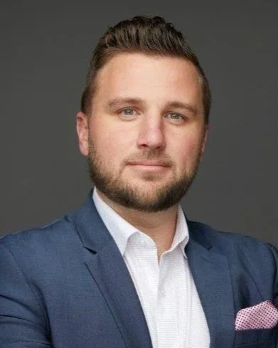 Headshot of a young man with light skin, dark brown hair, and a beard, wearing a navy blazer and white shirt, against a gray background.