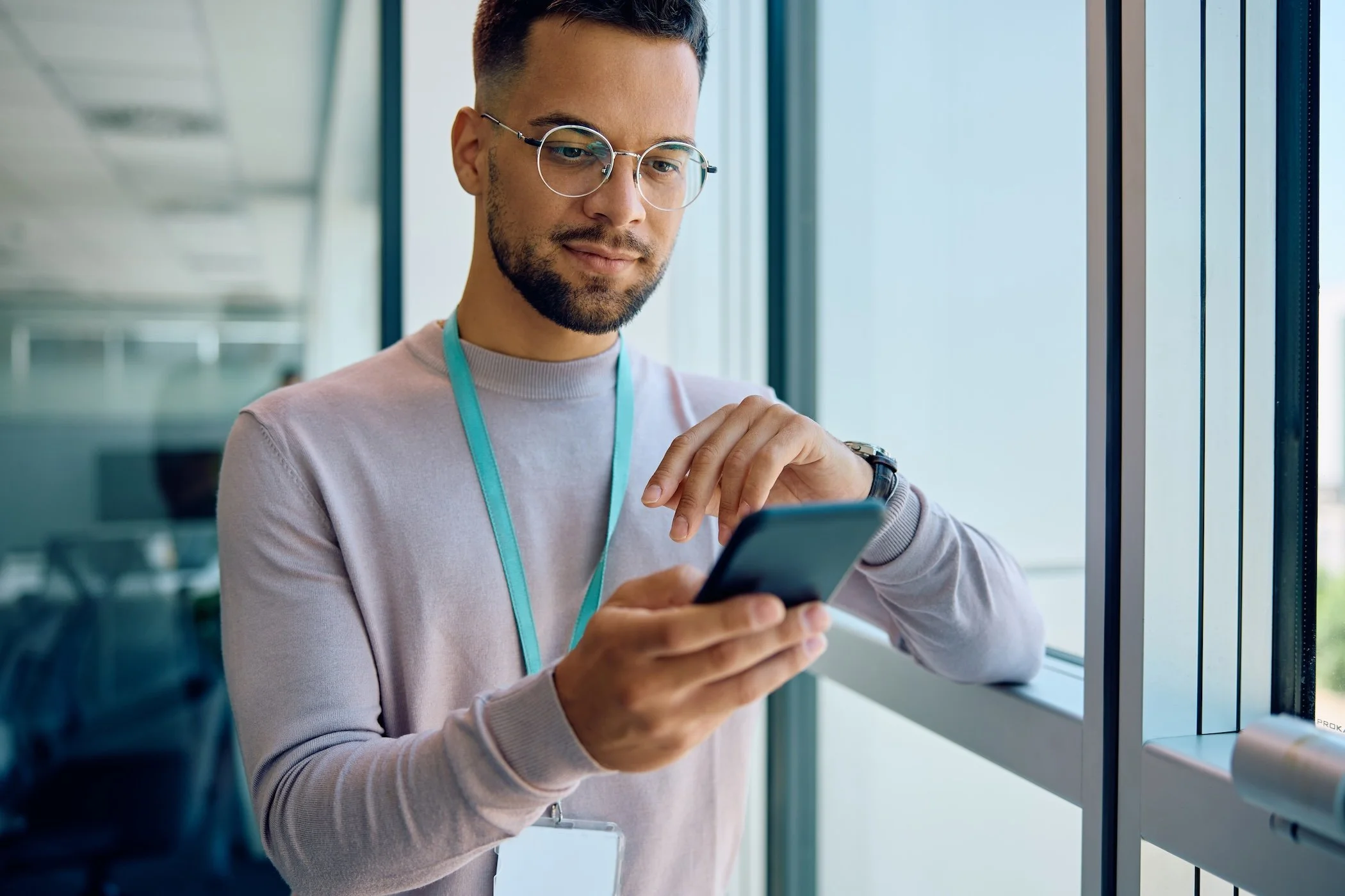 A man with glasses and a beard looking at his smartphone while standing by a window in an office building.