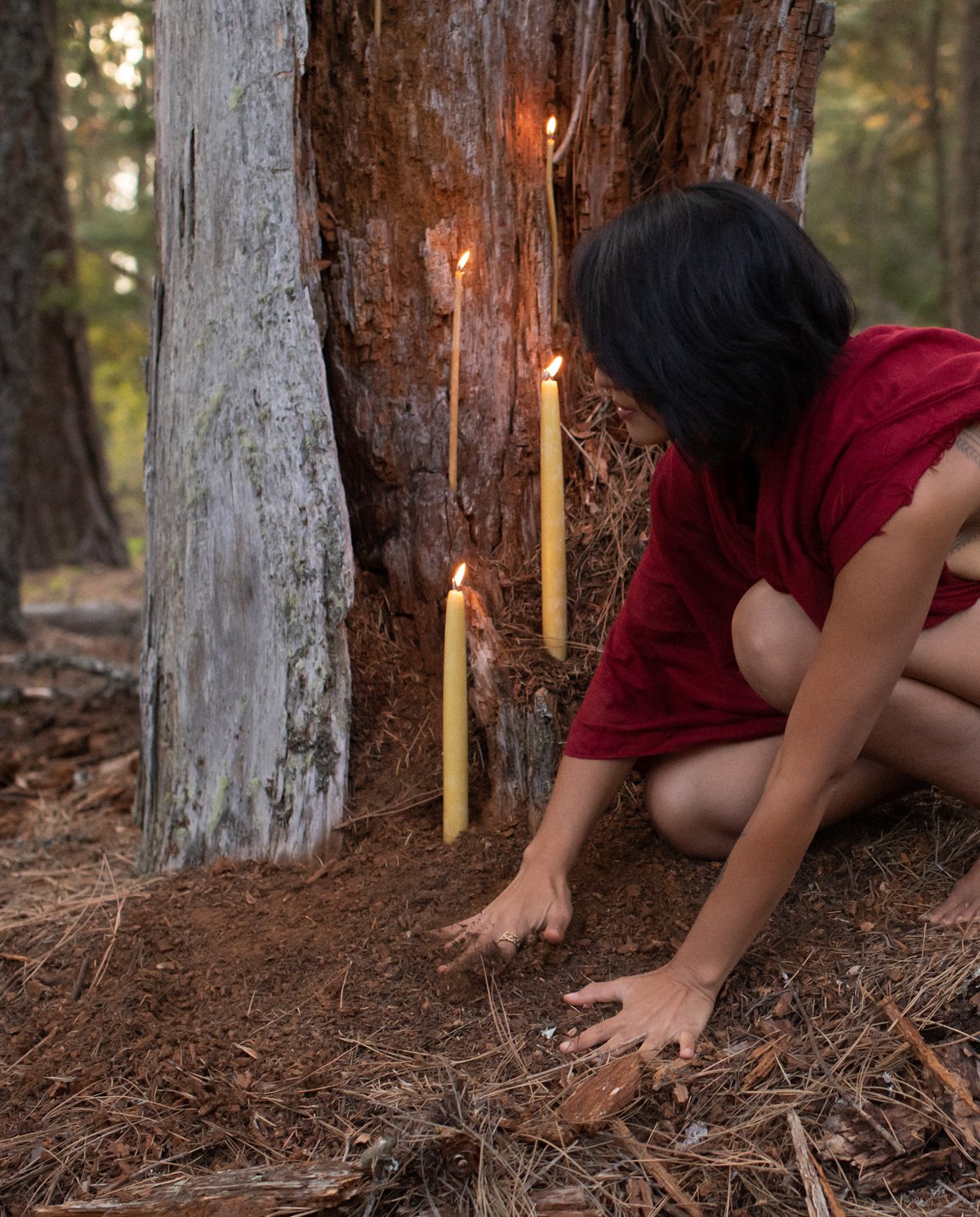 A person in a red dress kneels on the ground planting a seed in the soil next to a tree, with three lit candles placed vertically in the ground nearby, in a forest setting.