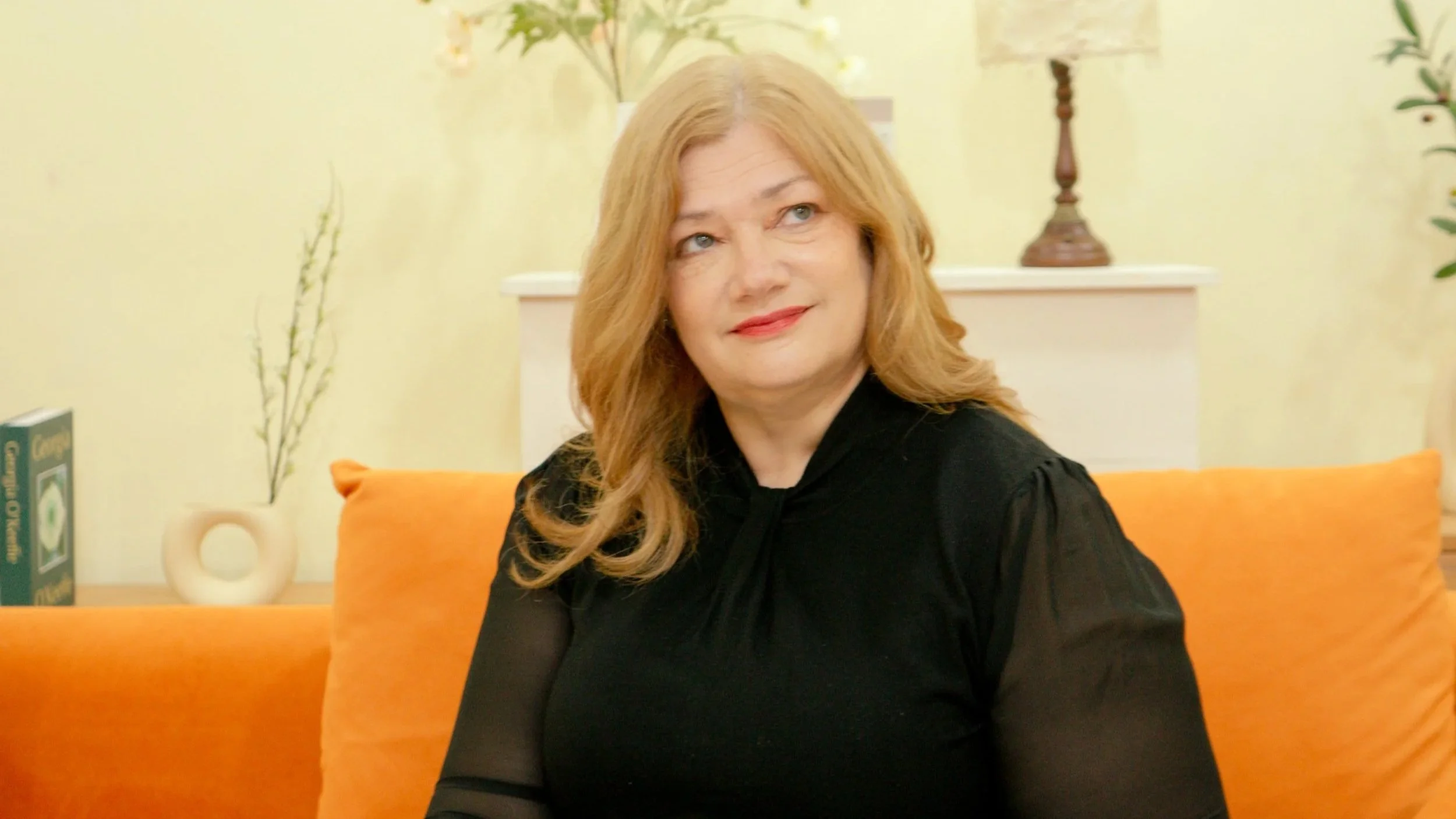 A woman with long red hair and a black top sits on an orange couch in a living room with light-colored walls, a white mantle with decorative items, and a book.