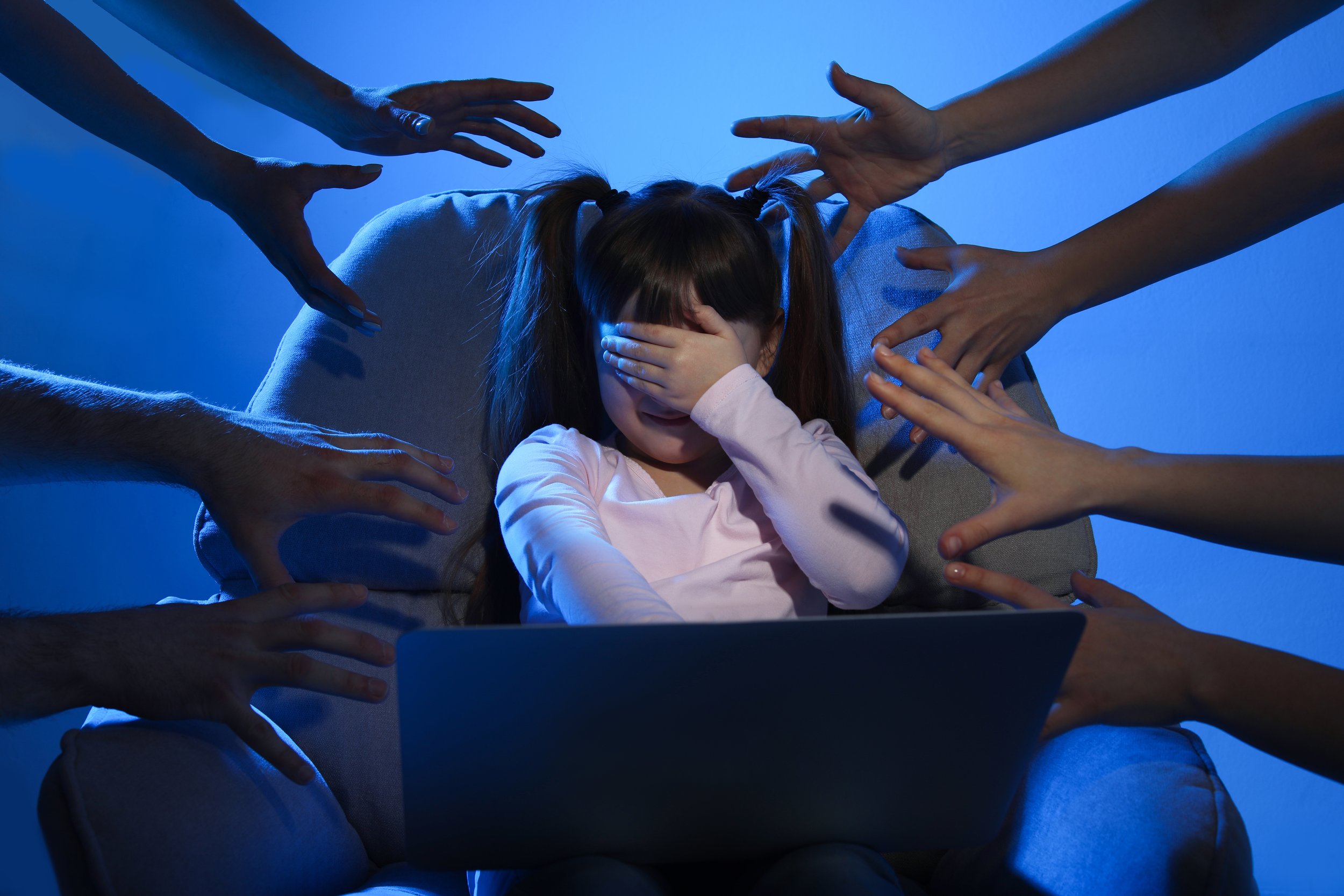 A young girl with pigtails covering her eyes and face, sitting on a couch with her hand over her face, surrounded by multiple hands reaching towards her in a dark blue lit room.
