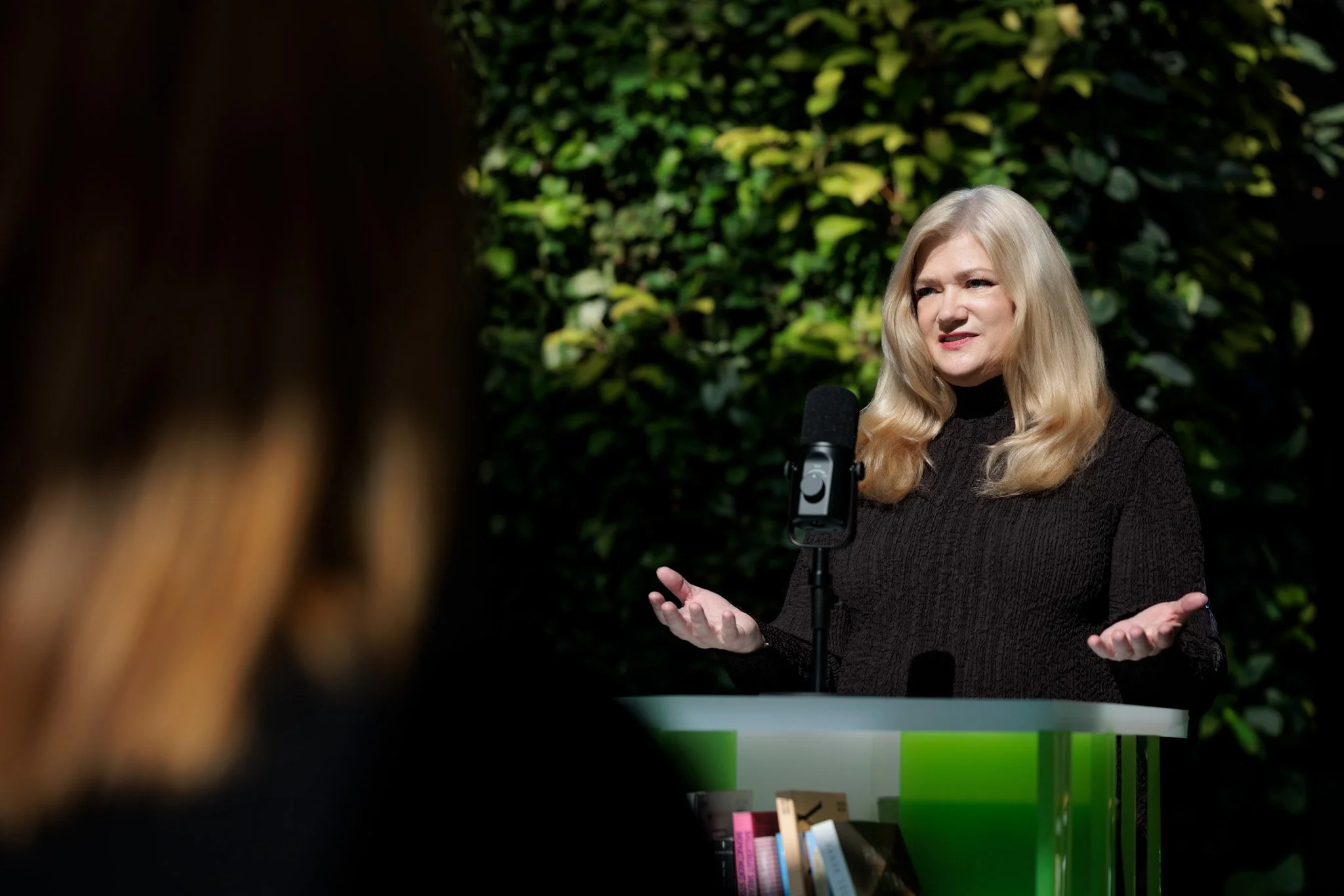 A woman with blonde hair in a black top speaking at a podium outdoors, with a microphone, and books on the podium, against a background of green foliage.