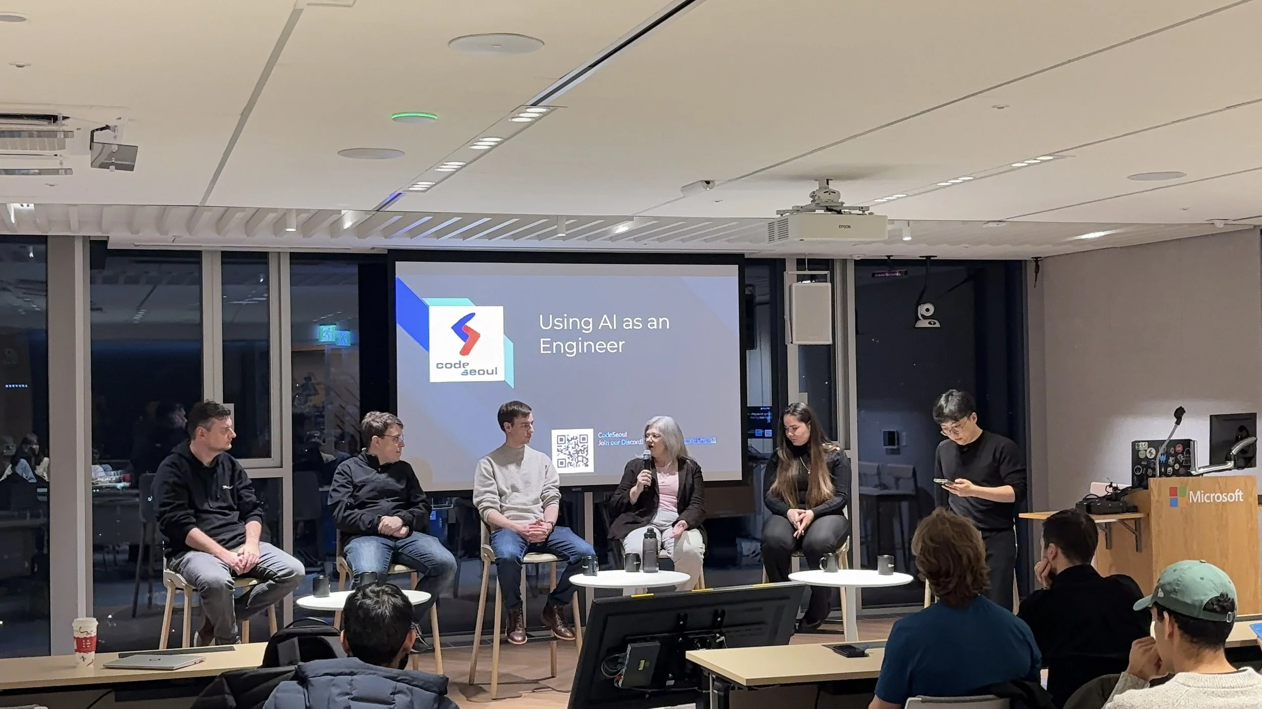 Panel discussion at a coding event titled "Using AI as an Engineer" with five people seated on stage in front of a screen displaying the event title and logo, audience members watching attentively, in a modern conference room.