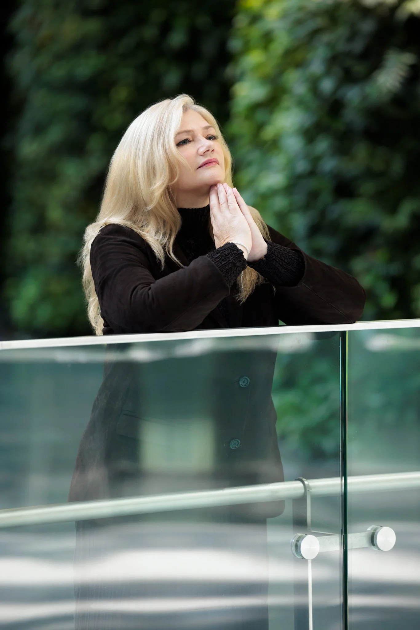 A woman with long blonde hair, dressed in a black coat, stands with her hands pressed together near her chin, looking contemplative against a blurred green outdoor background.