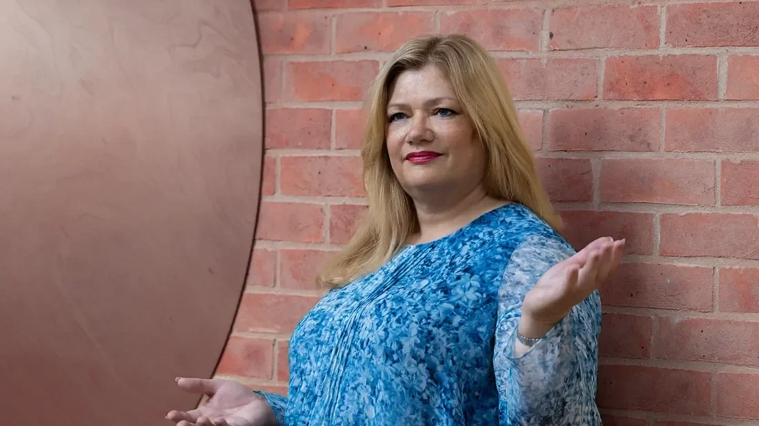 A woman with blonde hair wearing a blue patterned top, standing against a brick wall, with her hands raised in a shrugging gesture.