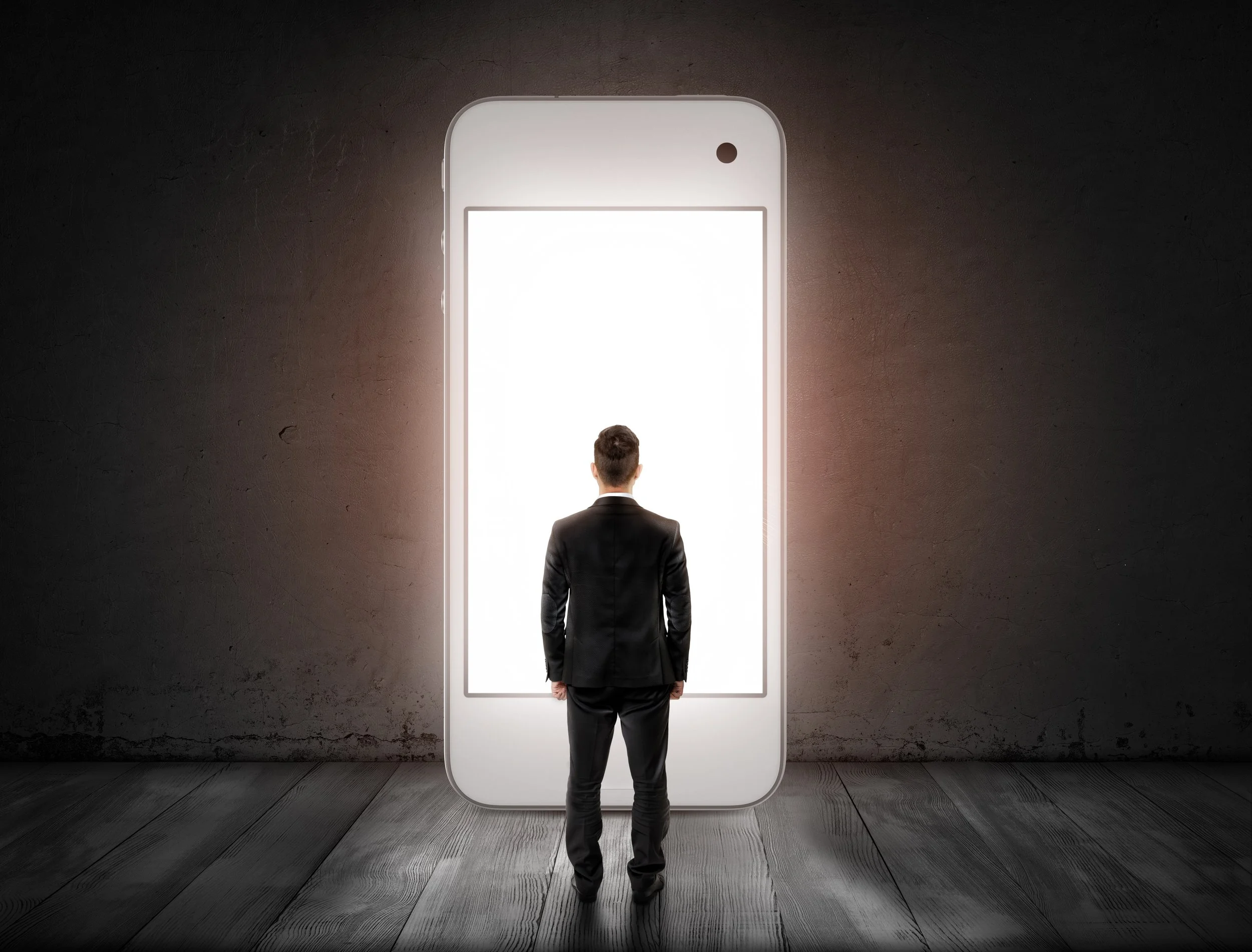 A man in a business suit stands facing a large smartphone screen mounted on a wall, with the screen showing a blank white background.
