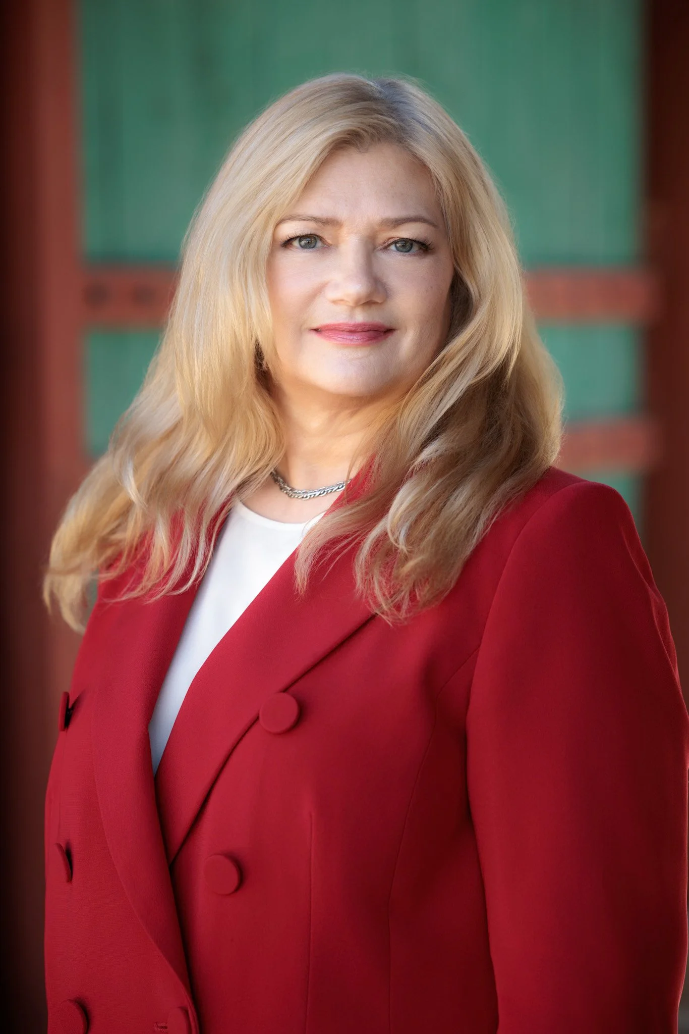 A woman with blonde hair wearing a red blazer and white top, standing outdoors with a green and red background.