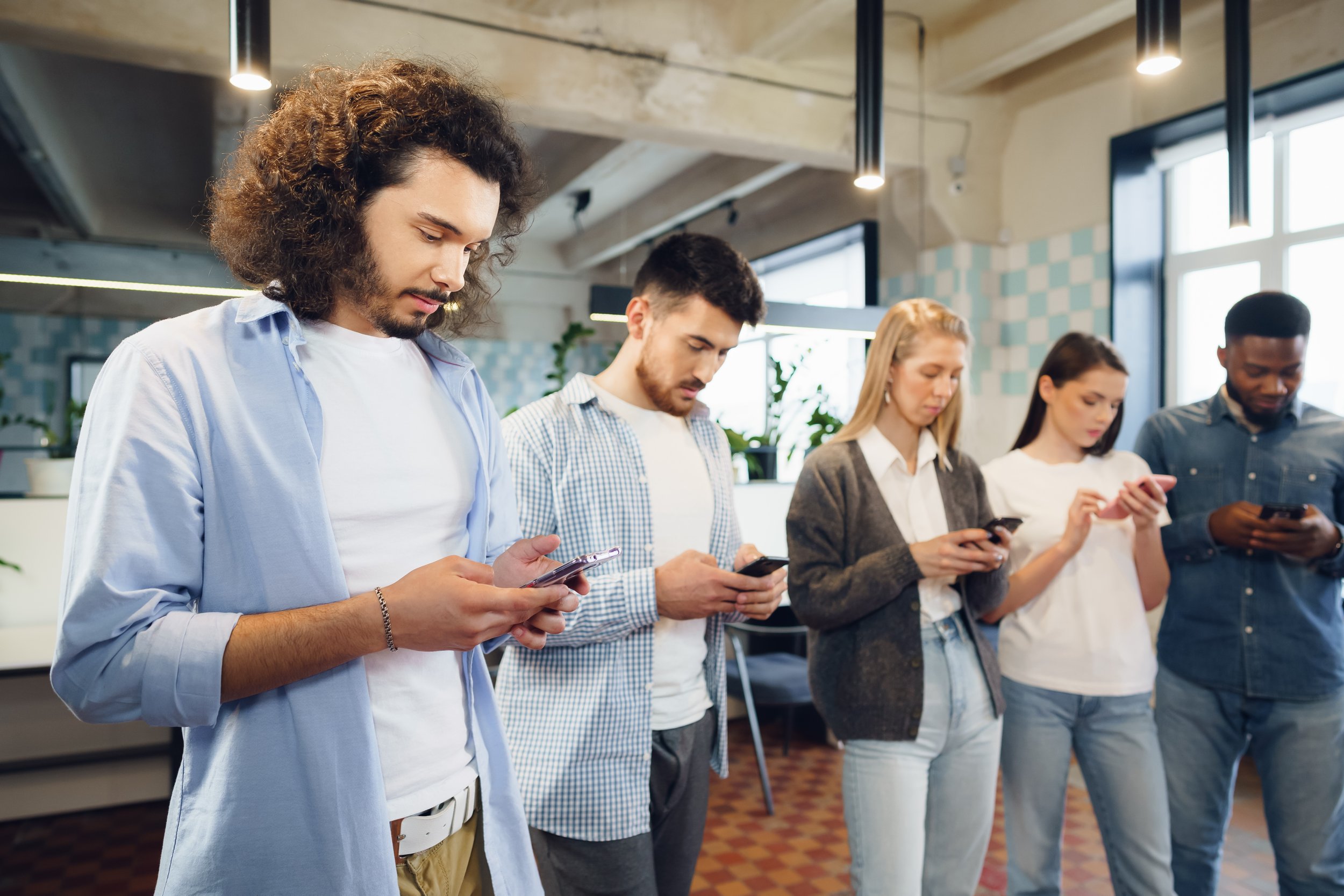 Five young adults standing in a row indoors, all looking at their smartphones, with a modern, well-lit background.