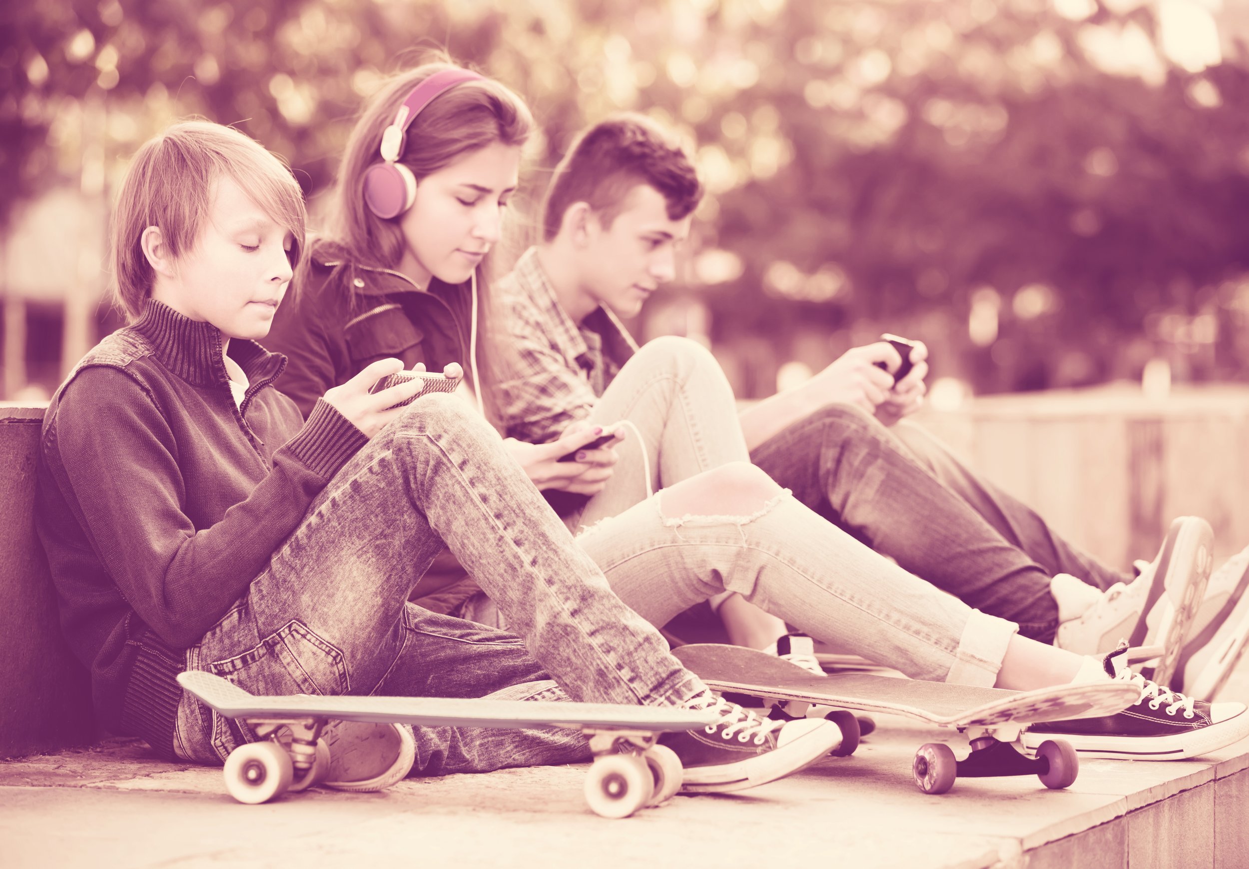 Three teenagers sitting on a bench outdoors, looking at their smartphones with skateboards and skate shoes next to them.