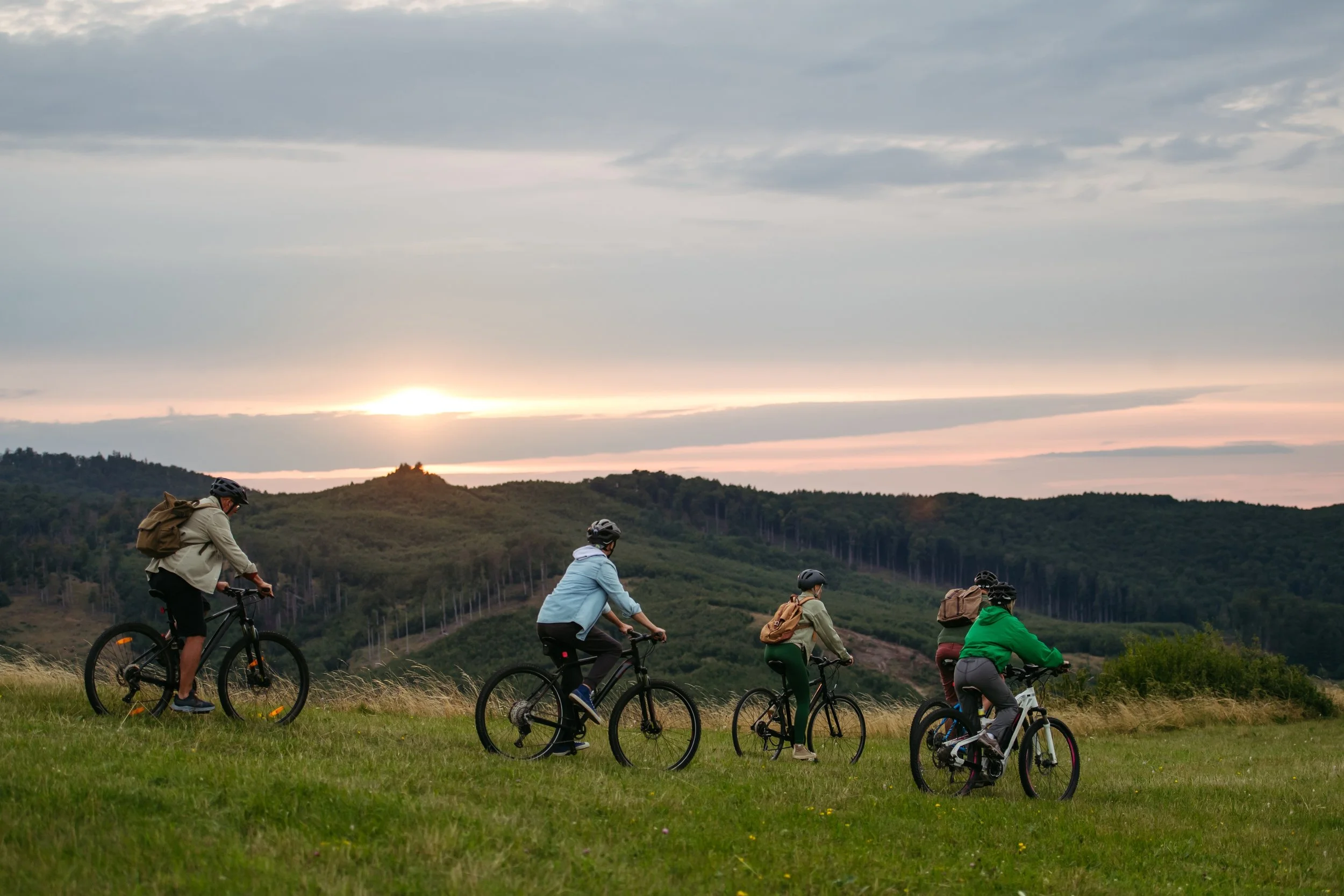 Five people riding bicycles on grassy terrain during sunset with hills and trees in the background.