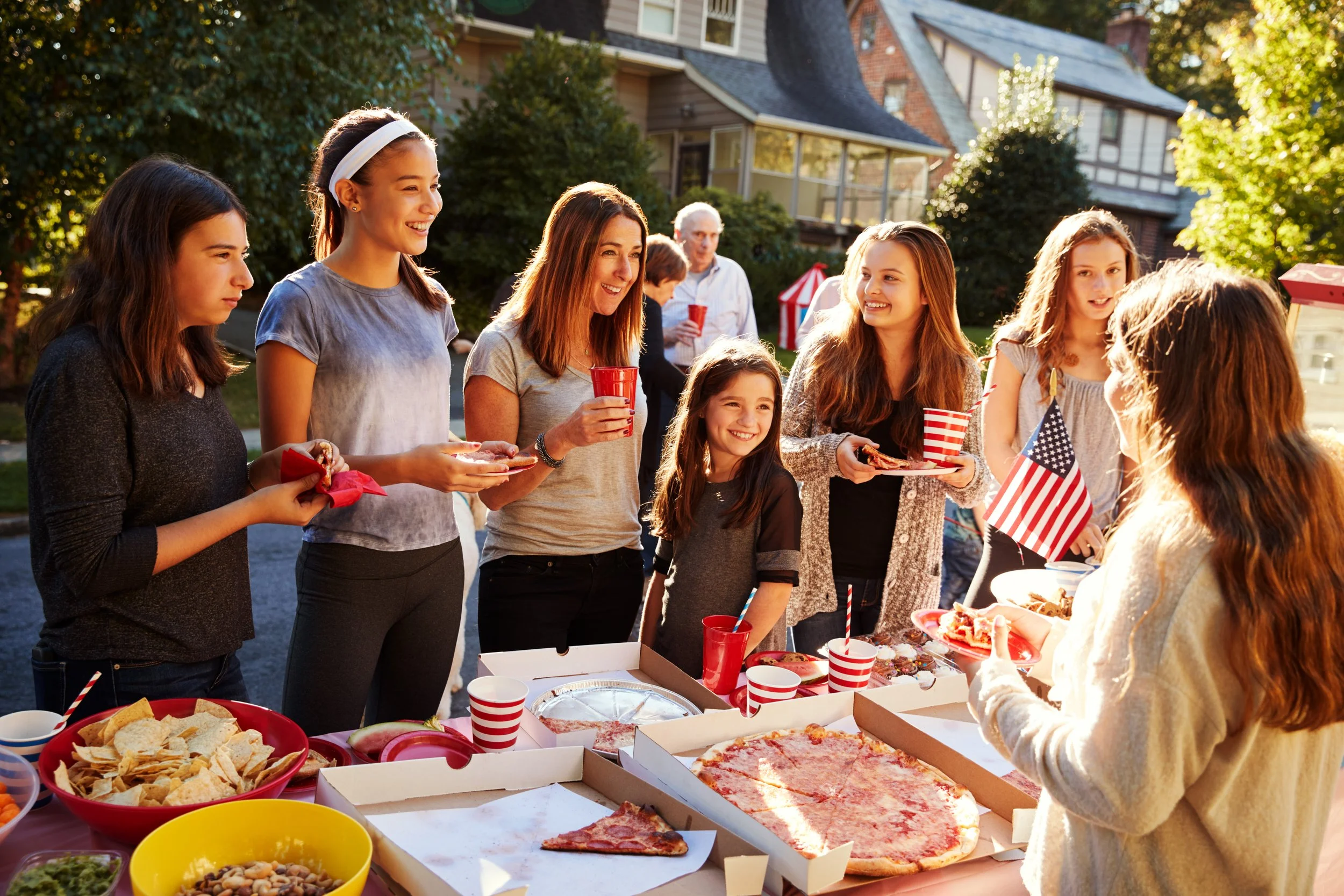 People gathered outside for a celebration with pizza, snacks, and drinks, with an American flag visible.