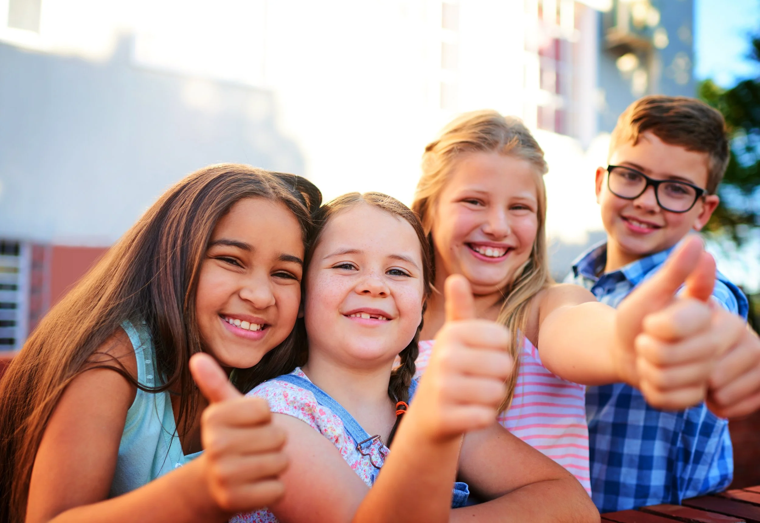 Four smiling children, two girls and two boys, giving thumbs up outside during daytime.