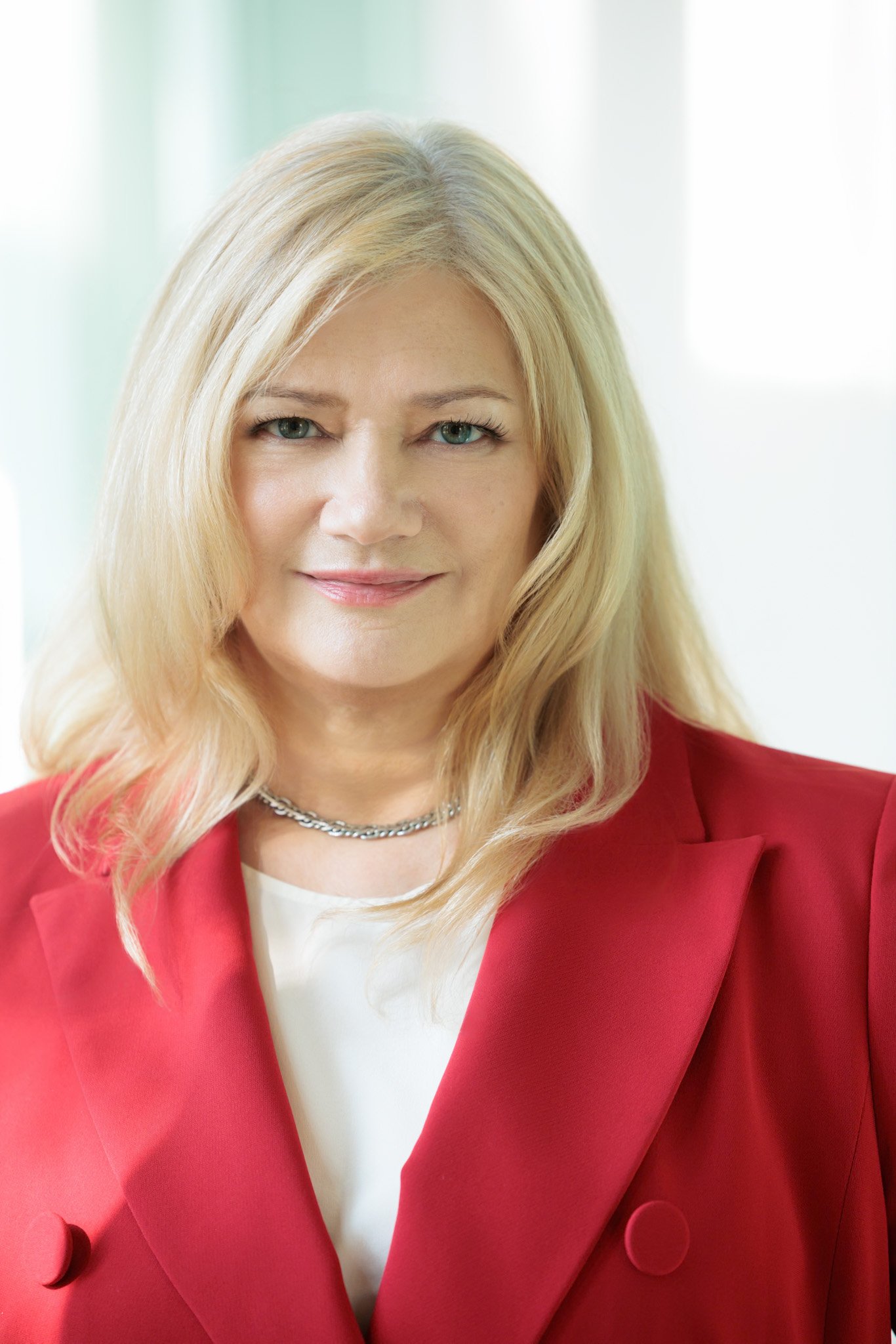 Portrait of a middle-aged woman with blonde hair wearing a red blazer and silver necklace, smiling gently.