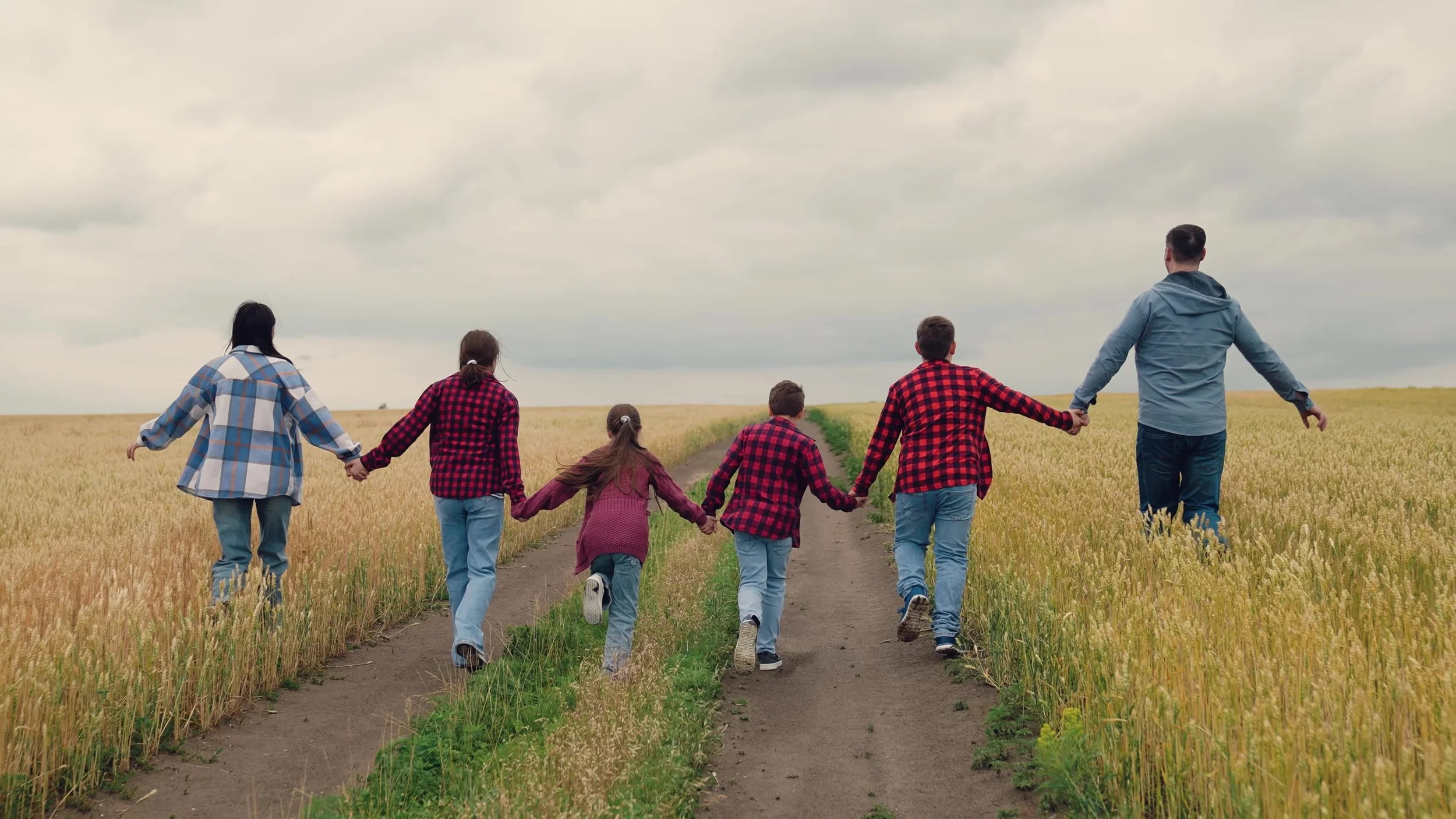 A family of six holding hands and walking along a dirt path through a wheat field under a cloudy sky.