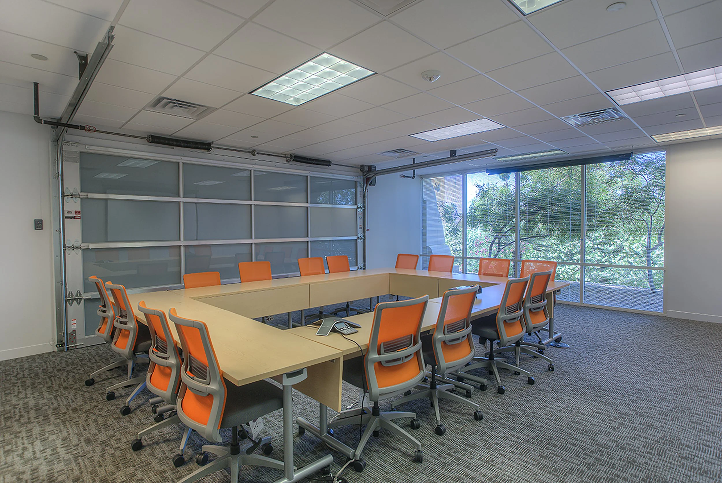 Empty conference room with orange chairs around a U-shaped table, a glass garage door, and large windows with trees outside.