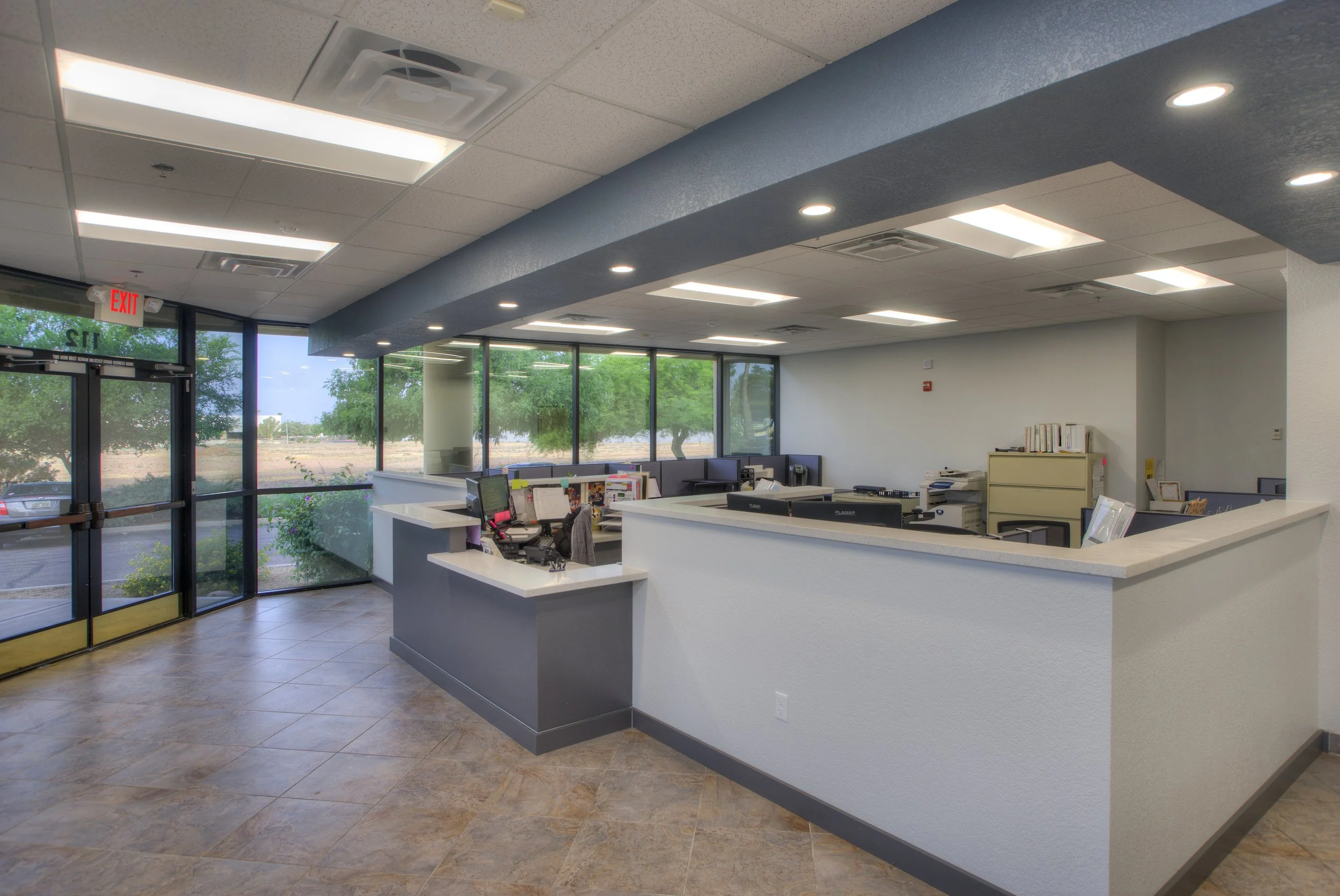 Empty office reception area with a curved counter, multiple desks with computers, placed in front of large windows showing greenery outside, and a glass entrance door with an exit sign above.