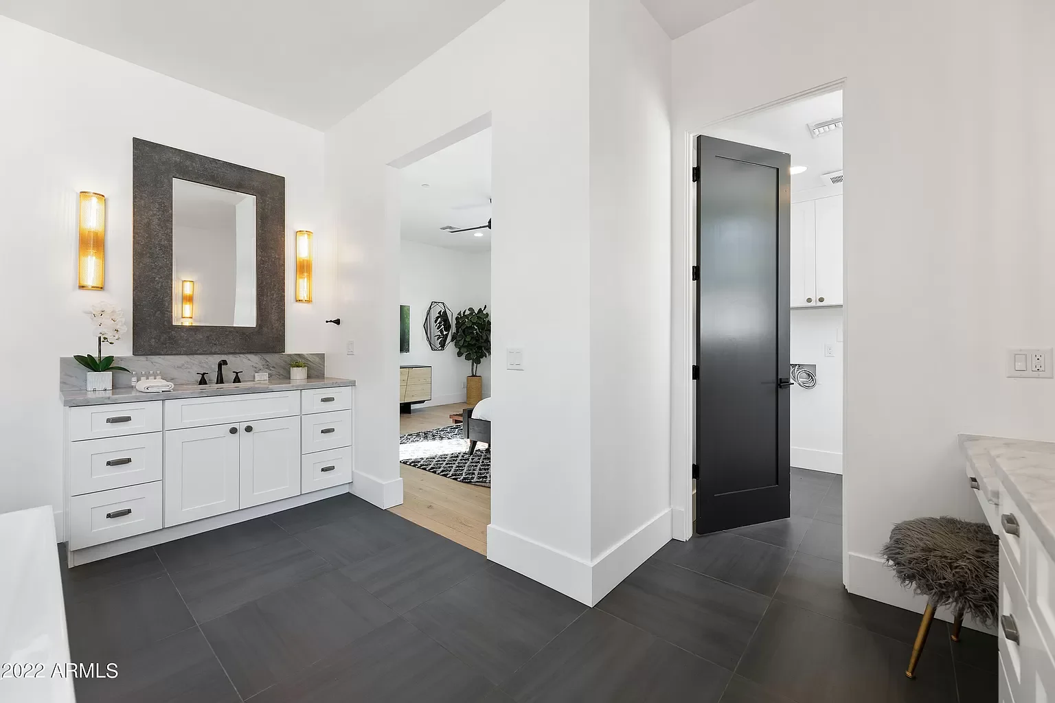 Modern bathroom with white cabinetry, marble countertop, black fixtures, and a large mirror surrounded by gold wall lights. The floor is dark tile, and there's a small furry stool on the right. An open door leads to a laundry area, with a glimpse int