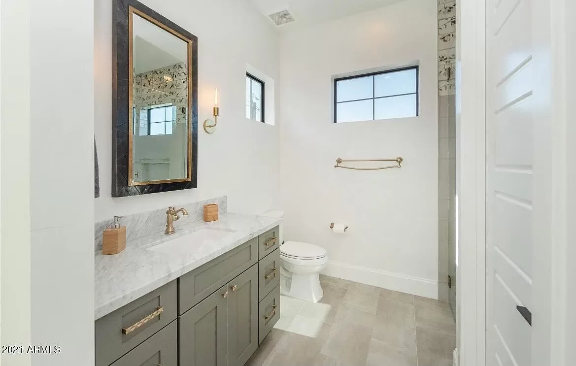 Modern bathroom with light gray vanity, white marble countertop, and gold hardware. Features a wall mirror and sconce light, small windows, and a toilet.