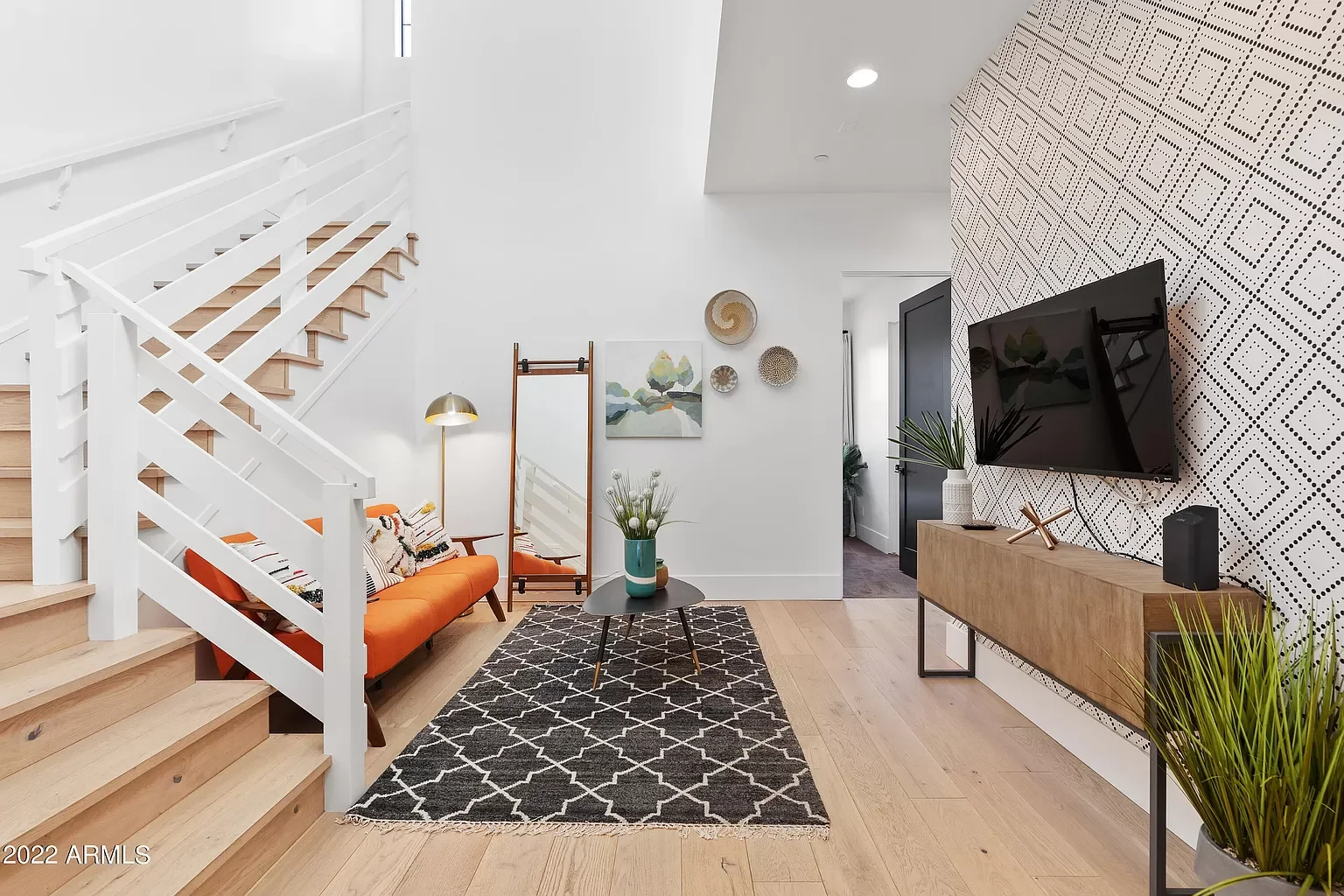 View of a modern living room with a white staircase, orange sofa, black and white rug, wall decor, a flat-screen TV, and wooden furniture, with a doorway leading to other rooms.
