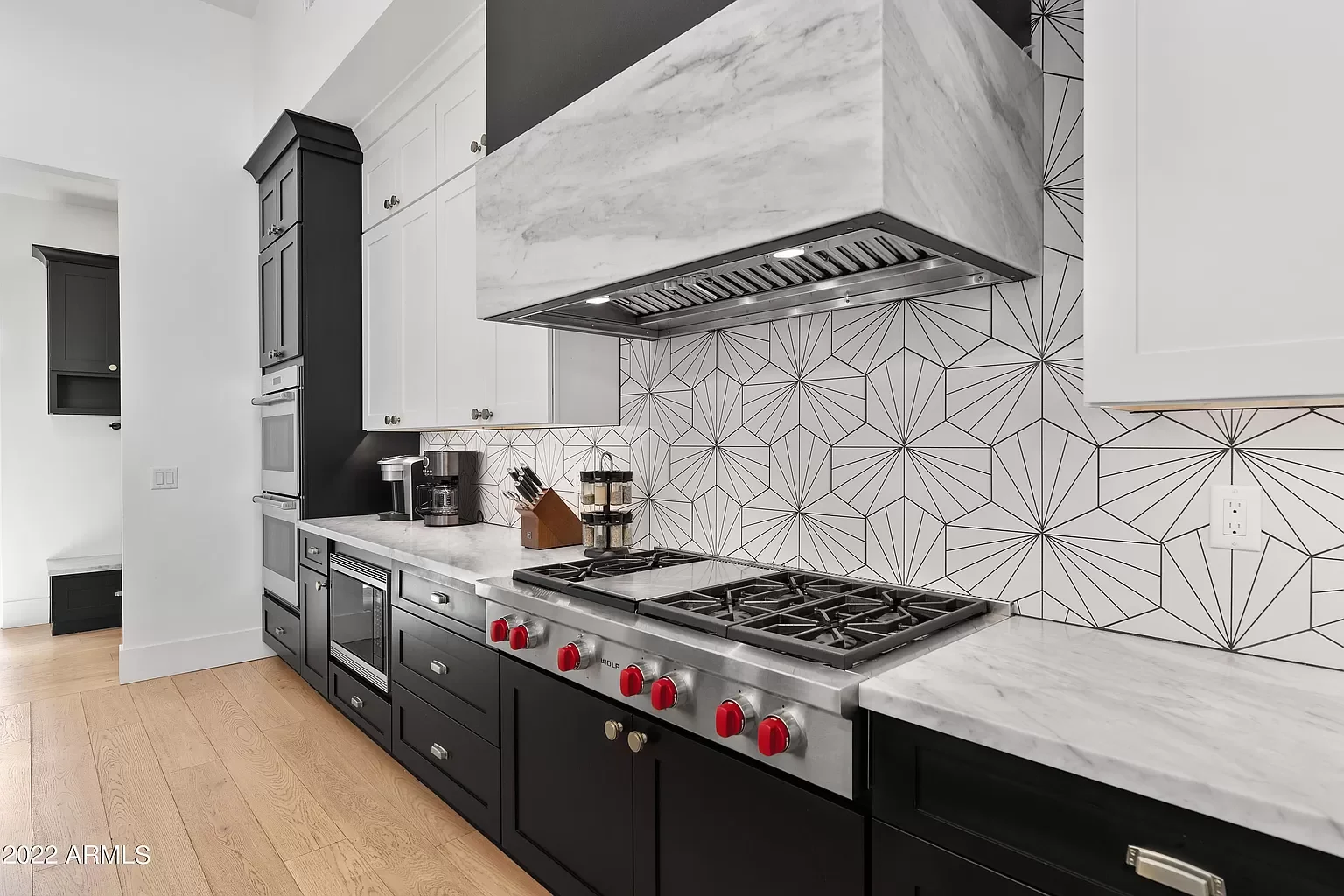 Modern kitchen with black cabinets, white marble countertops, geometric patterned backsplash, gas stove with red knobs, and built-in oven.