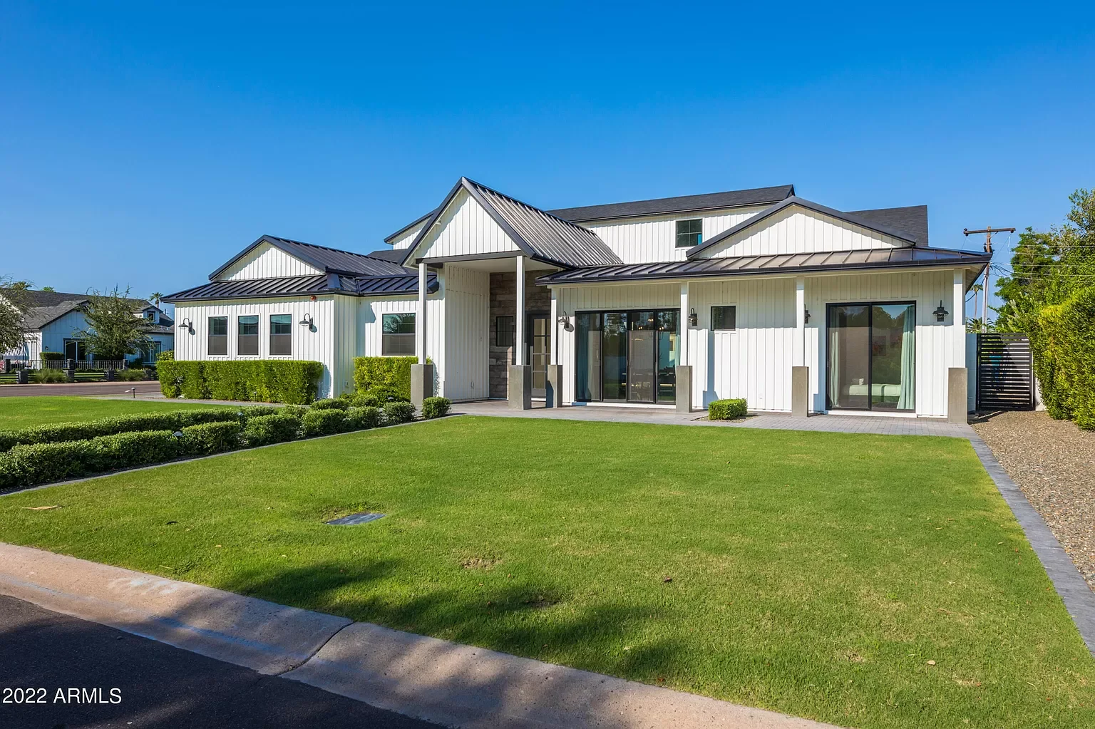 Modern two-story house with white vertical siding, metal roof, and large glass doors, surrounded by a well-manicured lawn and shrubbery under a clear blue sky.