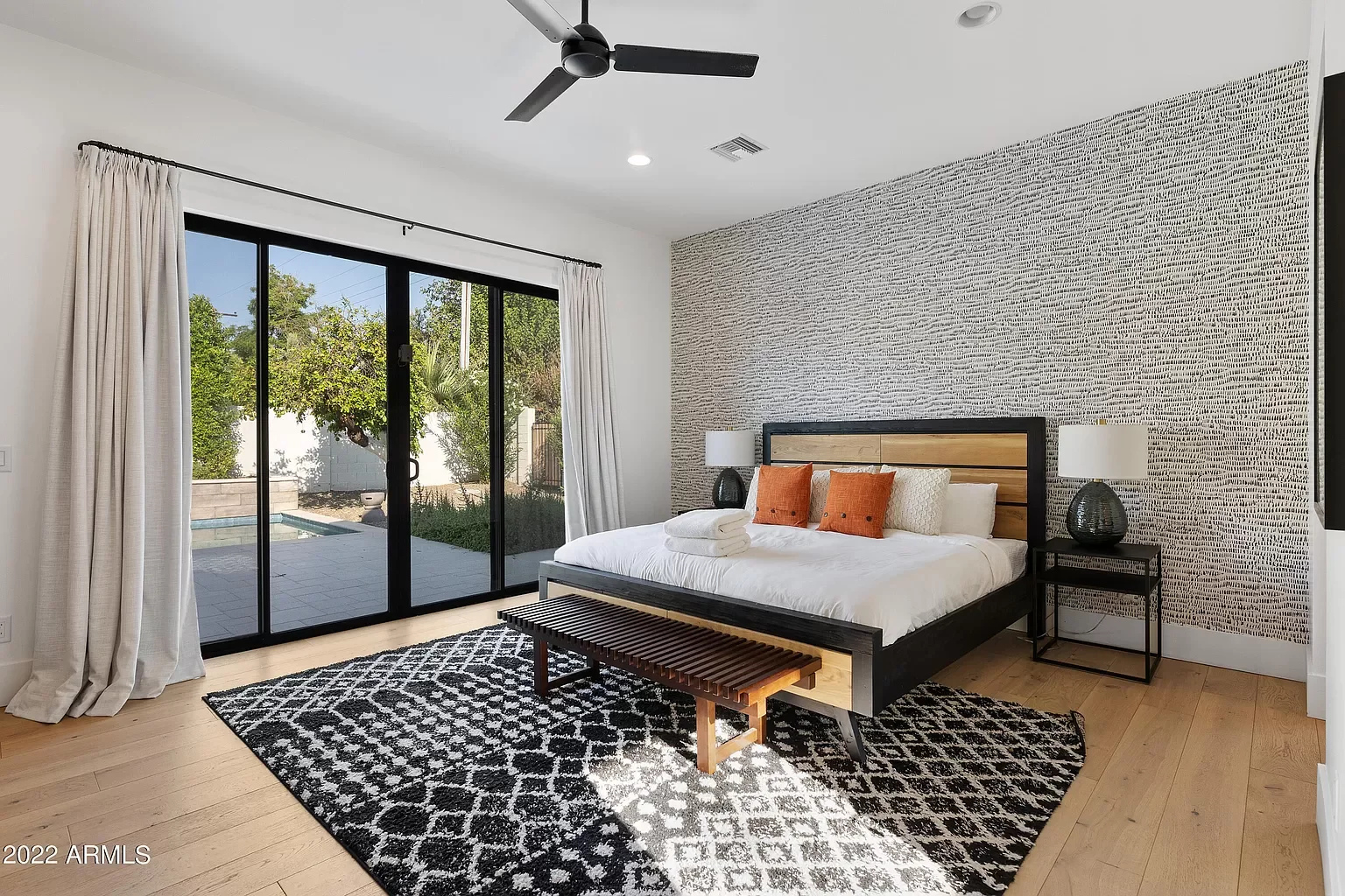 Modern bedroom with a king-sized bed, black frame, wooden headboard, orange throw pillows, white linens, white towel, black-and-white patterned rug, black table lamps, sliding glass door leading to backyard with patio, trees, and pool, beige curtains