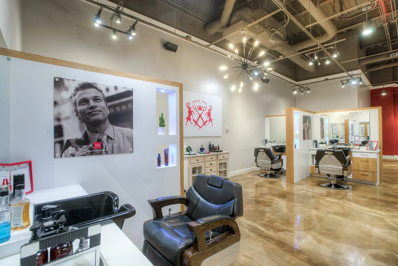 Modern barber shop with multiple styling stations, black leather chairs, large wall mirrors, wooden decor, and artwork on the wall, including a black-and-white portrait and a red emblem. The space has polished wooden floors and industrial ceiling lighting.