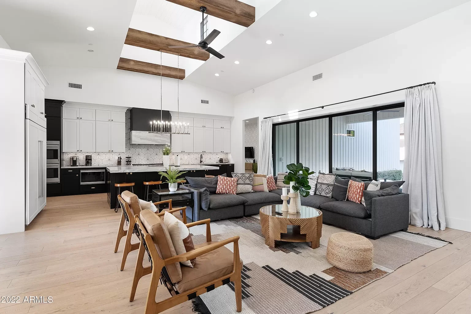 Open-concept living room and kitchen with high ceilings, large sliding glass doors, gray sectional sofa, wooden chairs, pendant lighting, and modern decor.