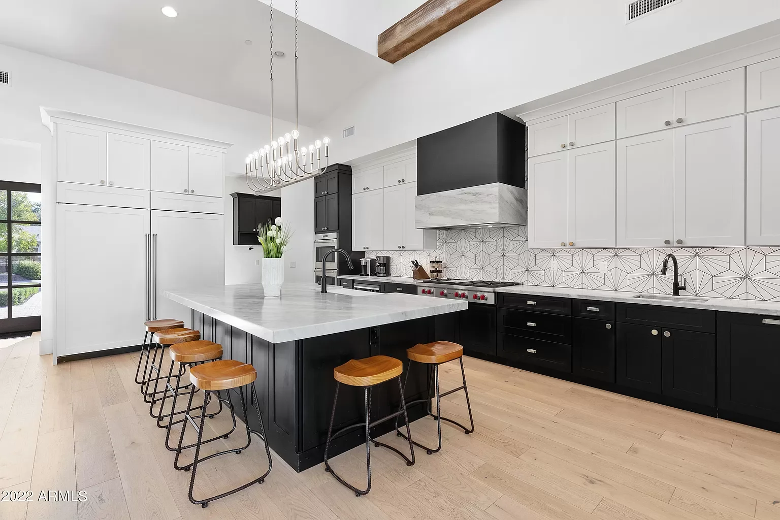 Modern kitchen with black and white cabinets, a large island with a white marble countertop, wooden stools, a geometric backsplash, stainless steel appliances, and a hanging chandelier.