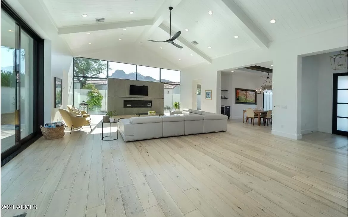 Bright, modern living room with white walls, light wood flooring, and large windows. Features a white sectional sofa, two yellow accent chairs, and a ceiling fan. In the background, there's a fireplace with a mounted TV above and a view of outdoors w