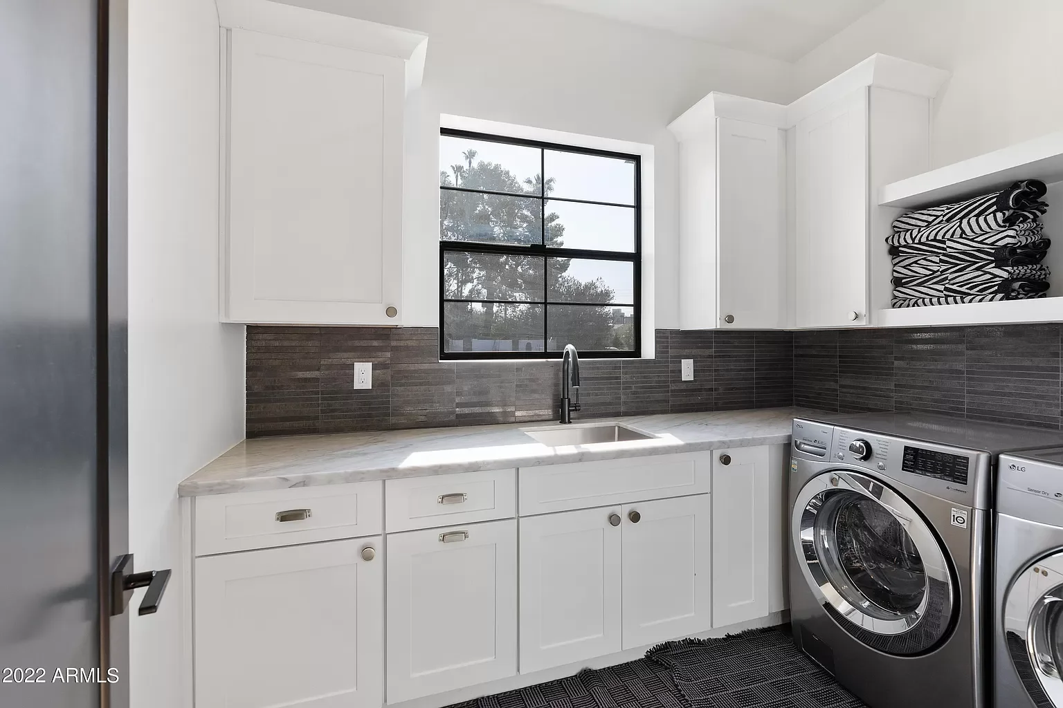 Laundry room with white cabinets, a marble countertop, a black sink faucet, a window with a view of trees, and a stacked set of black and white towels on open shelving. There are washer and dryer units.