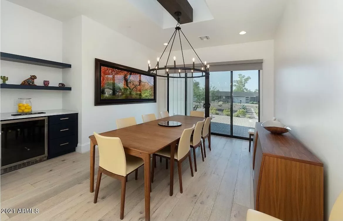 Modern dining room with a wooden table, eight cream-colored chairs, a black chandelier, and sliding glass doors leading outside. There are wall-mounted black shelves with decorative items and a sideboard on the right.